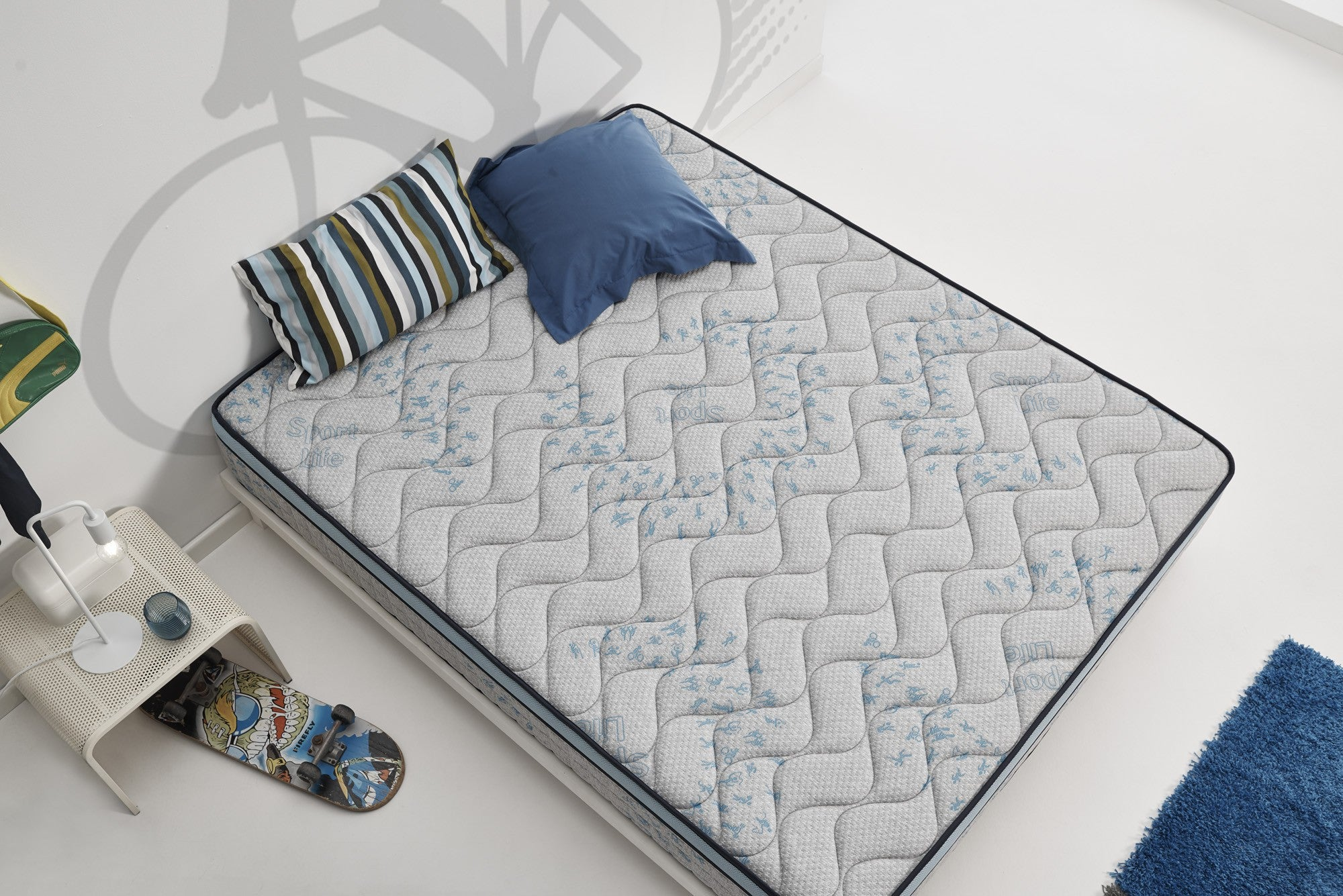 Una cama con almohadas y una patineta. Colores: blanco, azul y beige.