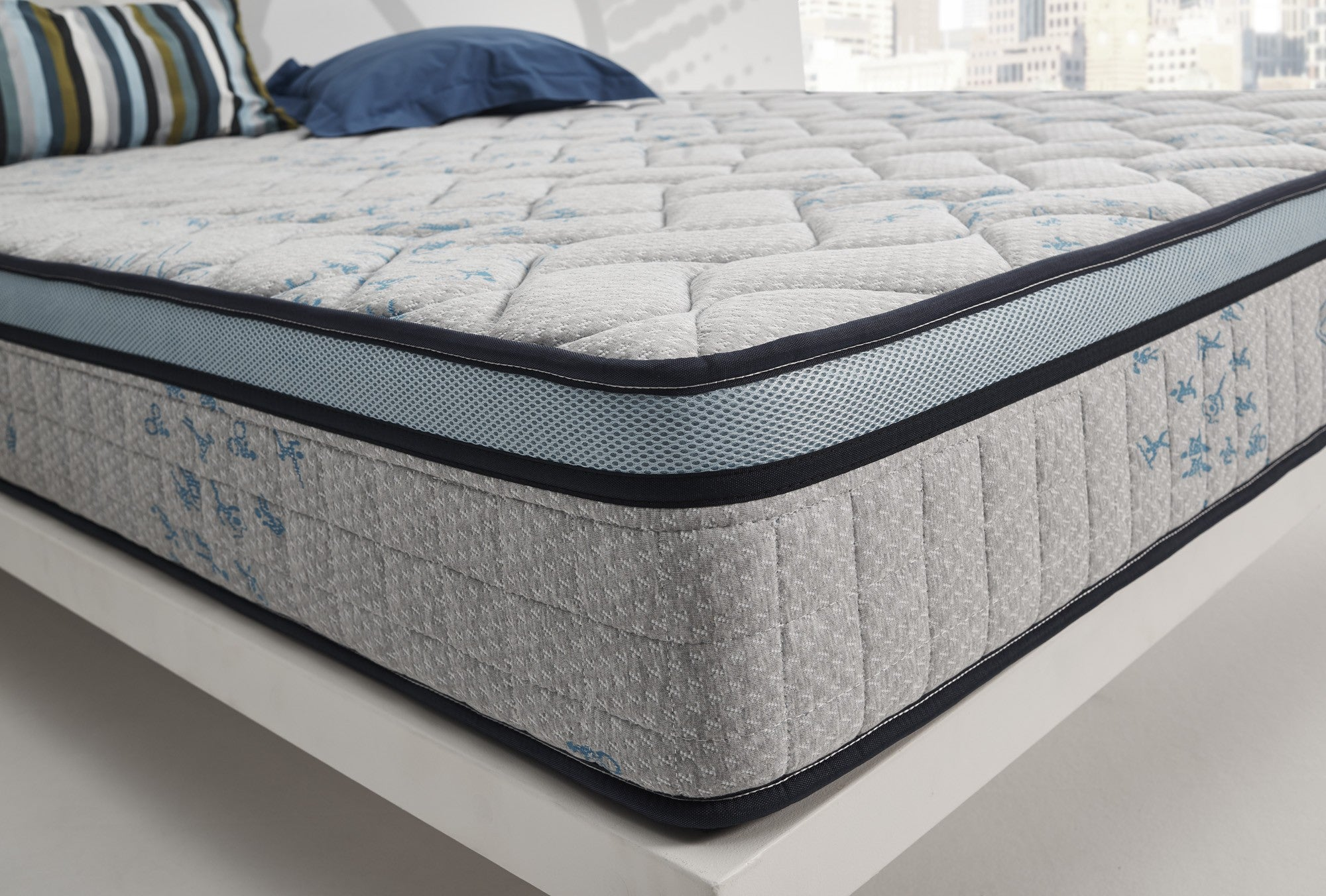 Un colchón sobre un marco de cama blanco. Las almohadas están encima. El fondo tiene una vista de la ciudad.