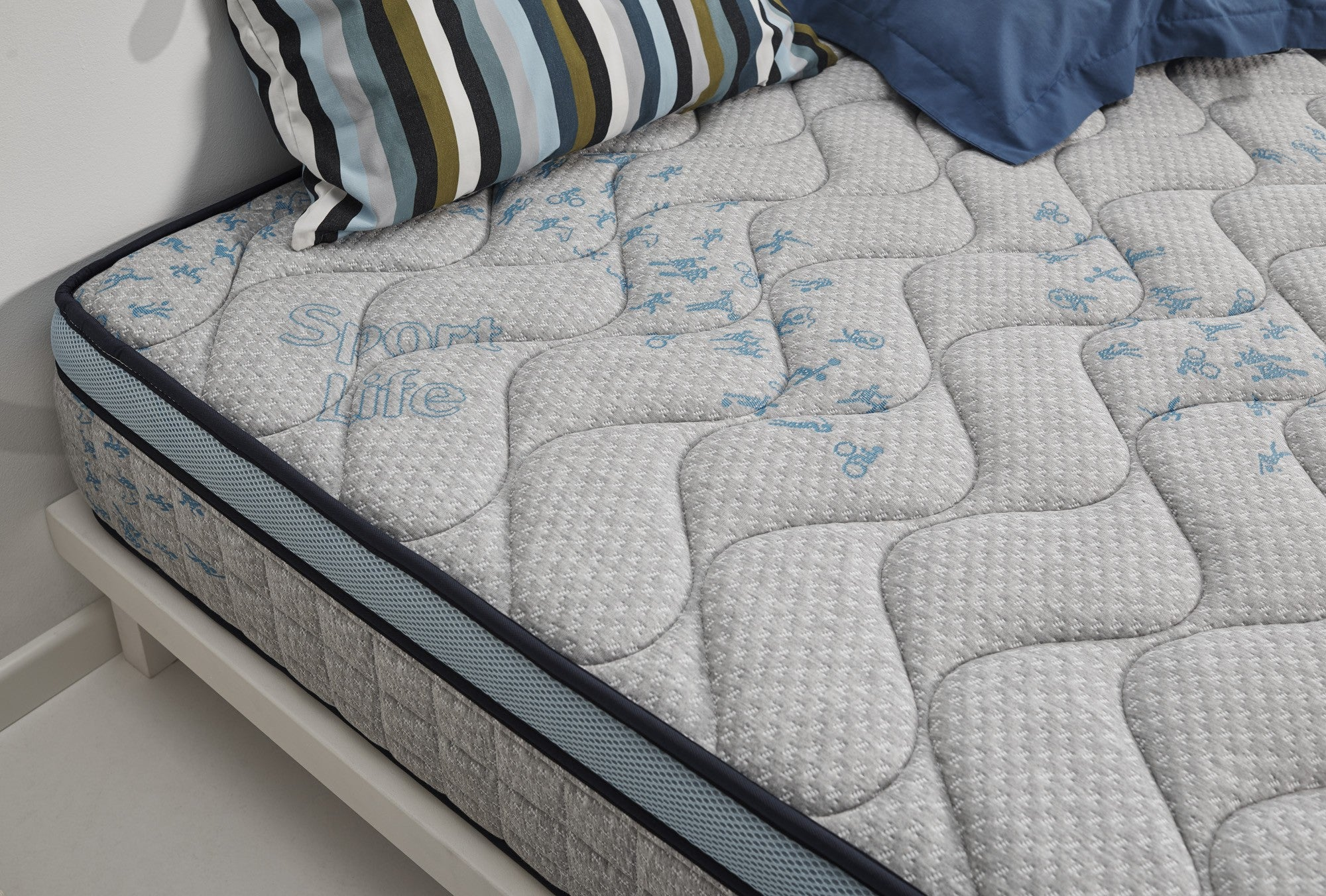 Colchón gris con detalles azules y almohadas. Almohada a rayas visible. Somier blanco.