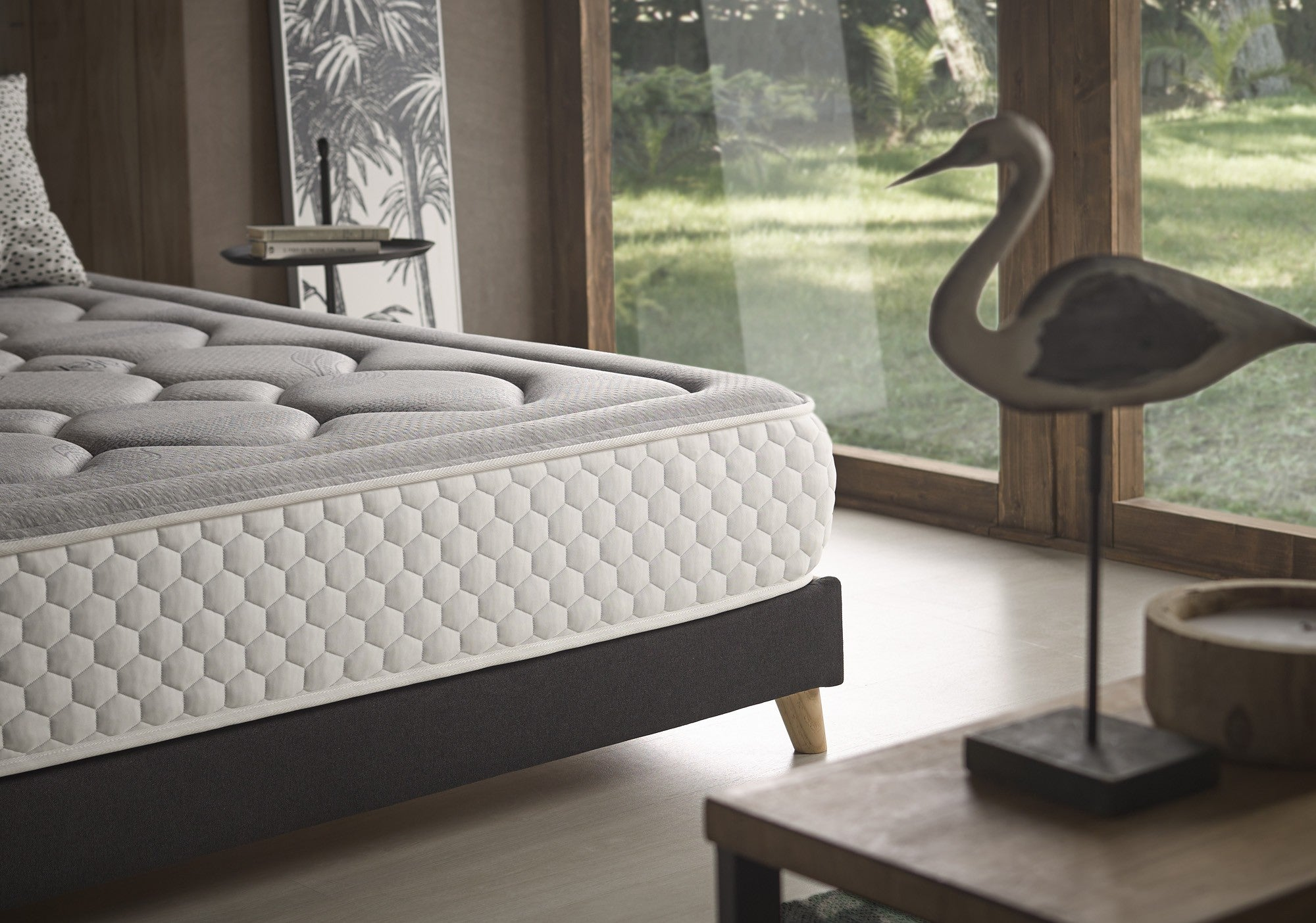 Primer plano de colchón gris en una cama con mesita de noche de madera y pájaro decorativo.