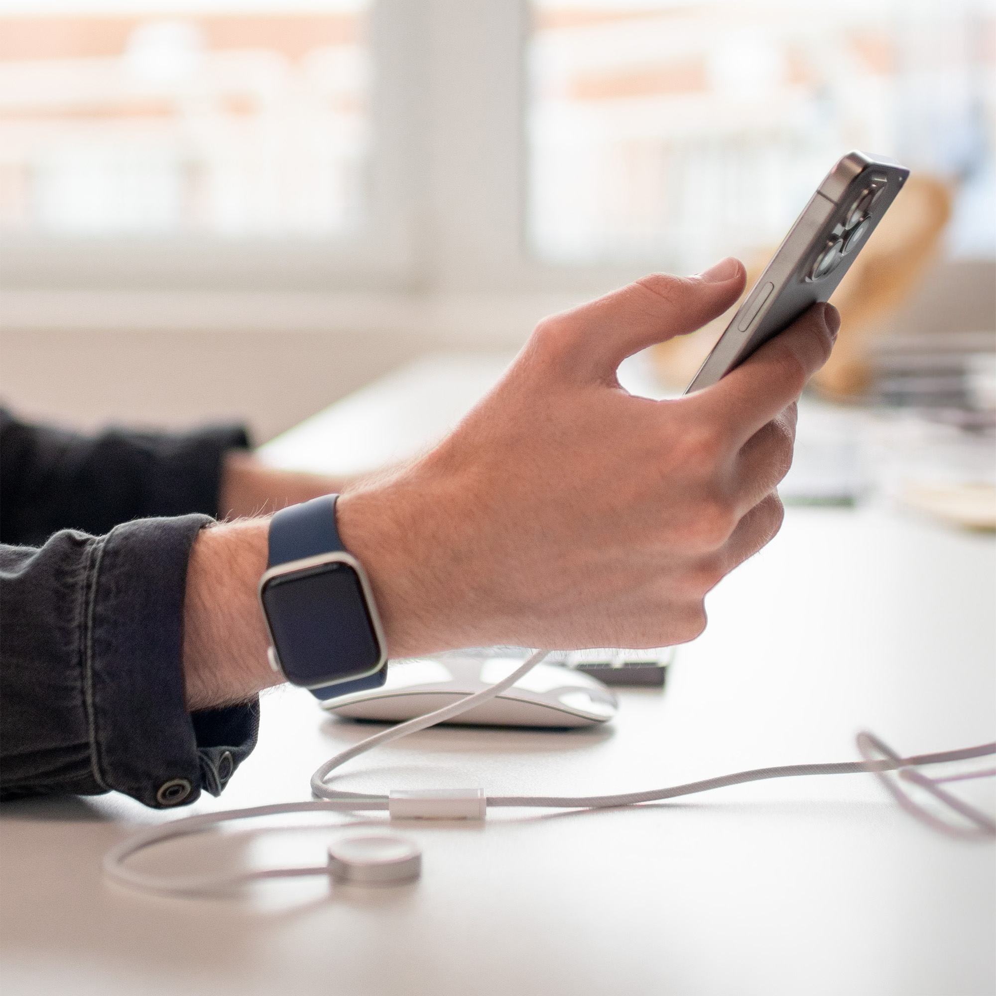 Eine Person hält ein Telefon und trägt eine Smartwatch. Weißer Schreibtisch mit Kabel und Maus. Verschwommener Hintergrund.