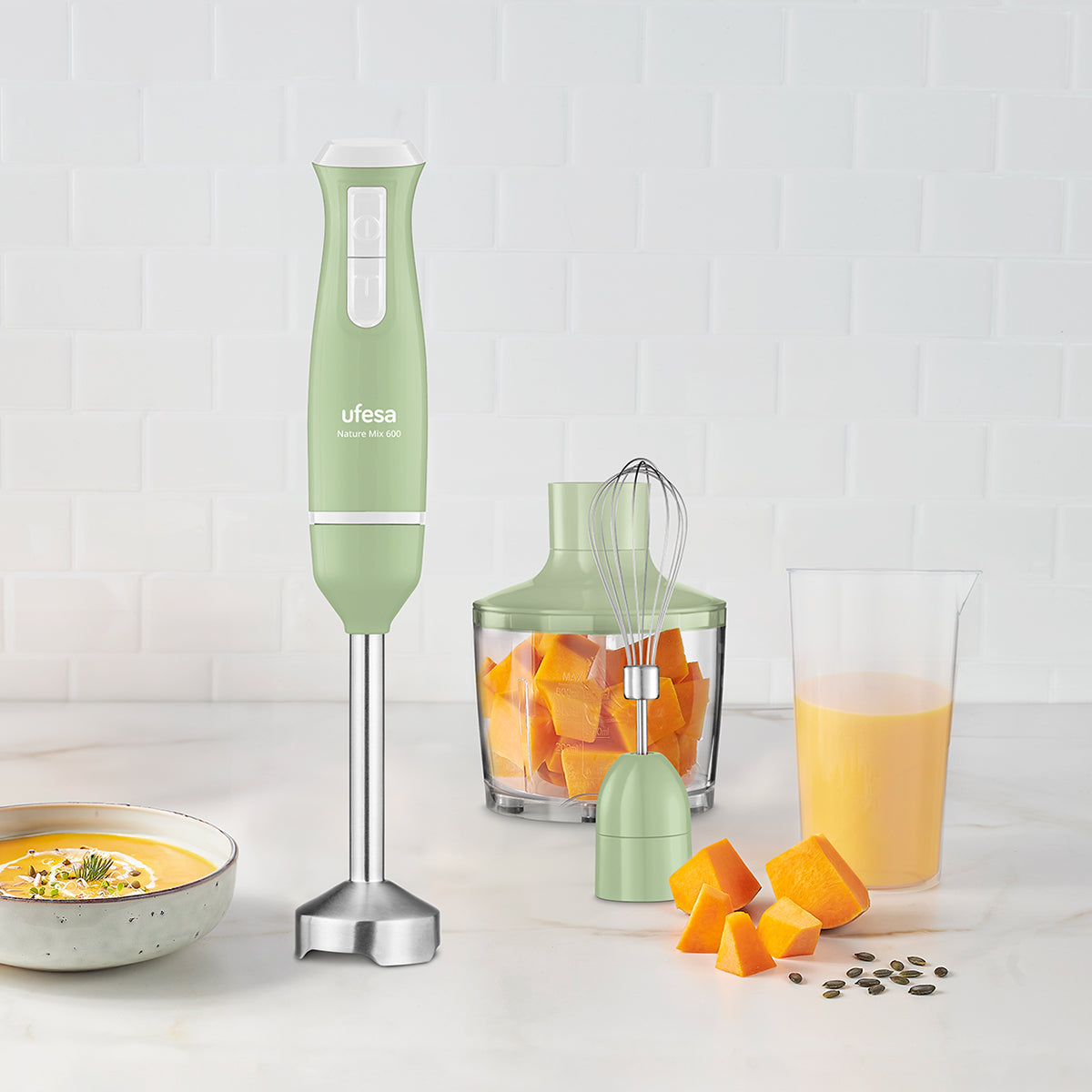 Batidora de mano con accesorios y trozos de calabaza sobre una superficie blanca.