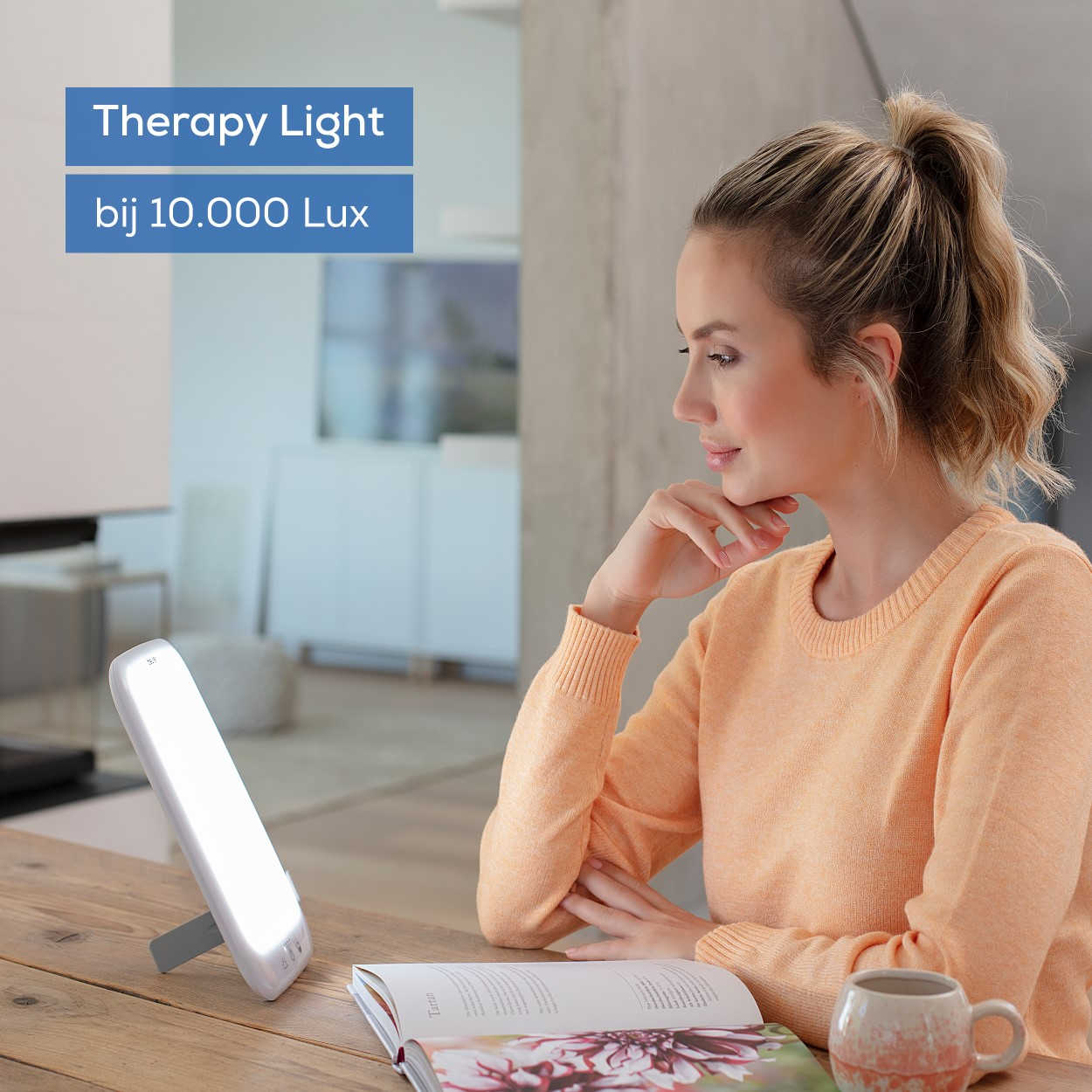 Eine Frau sitzt an einem Tisch und liest ein Buch, mit einer Therapielampe und einer Tasse.