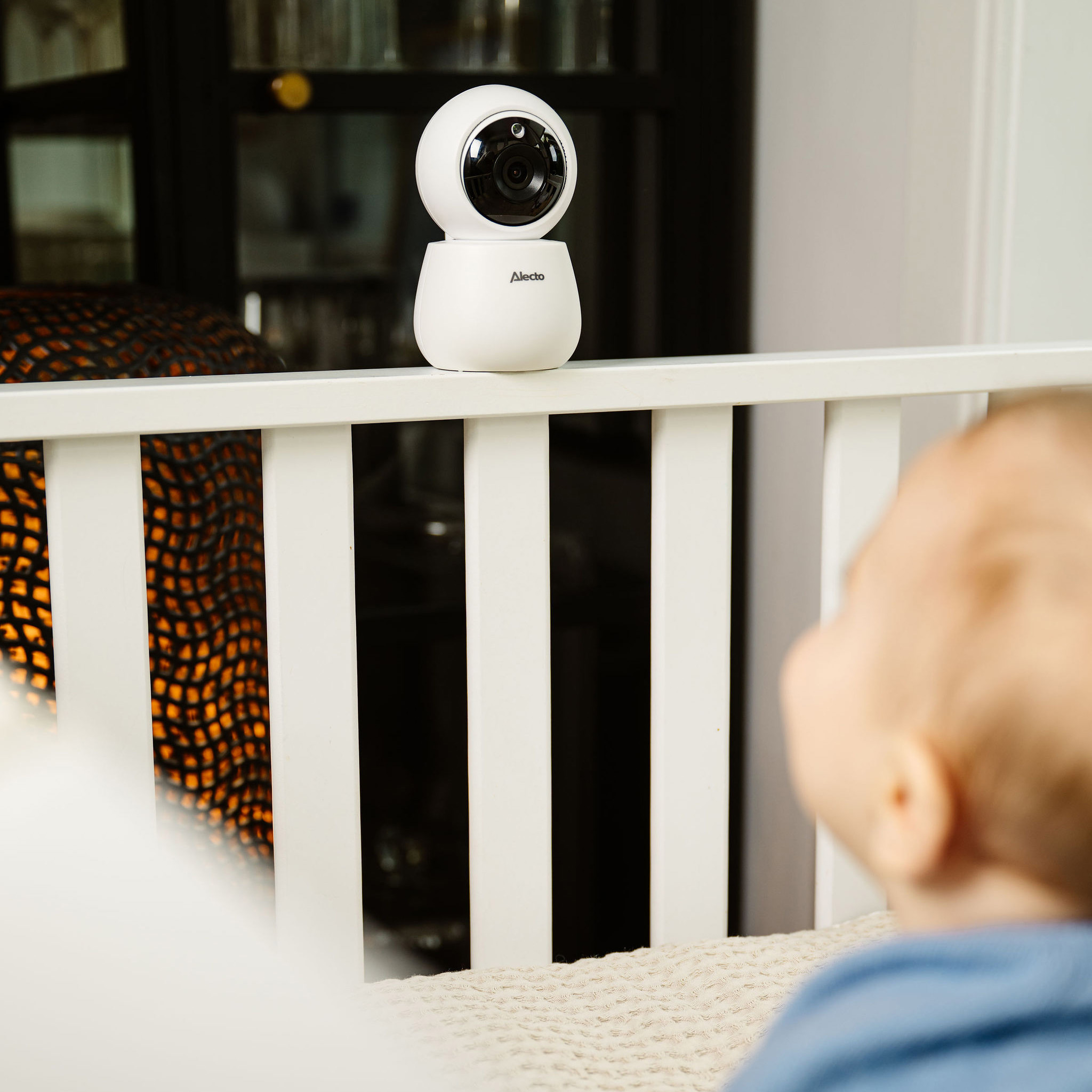 Eine weiße Babyfonkamera an einer weißen Krippenschiene. Ein Baby ist im Bett, unscharf.