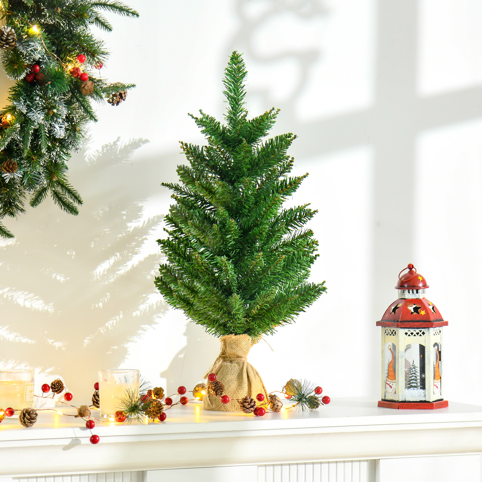 Pequeño árbol de Navidad artificial en arpillera, con adornos. Guirnalda y farol en superficie blanca.