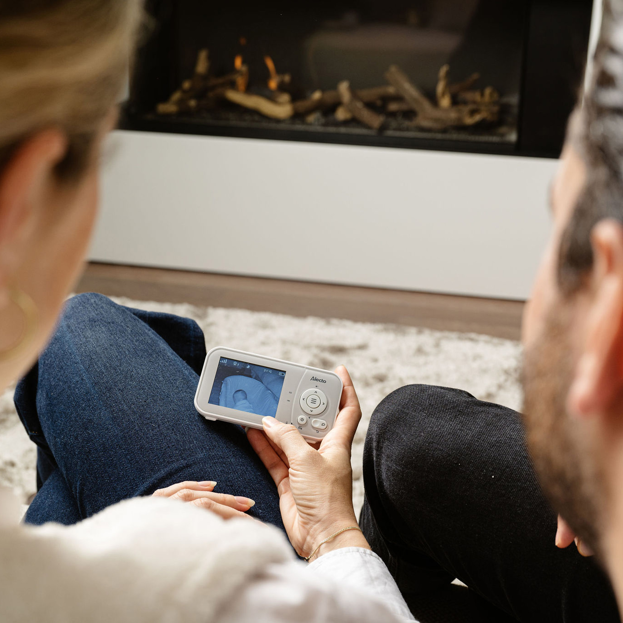 Ein Paar betrachtet einen Babyphone-Monitor. Sie sitzen auf einem Teppich vor einem Kamin.