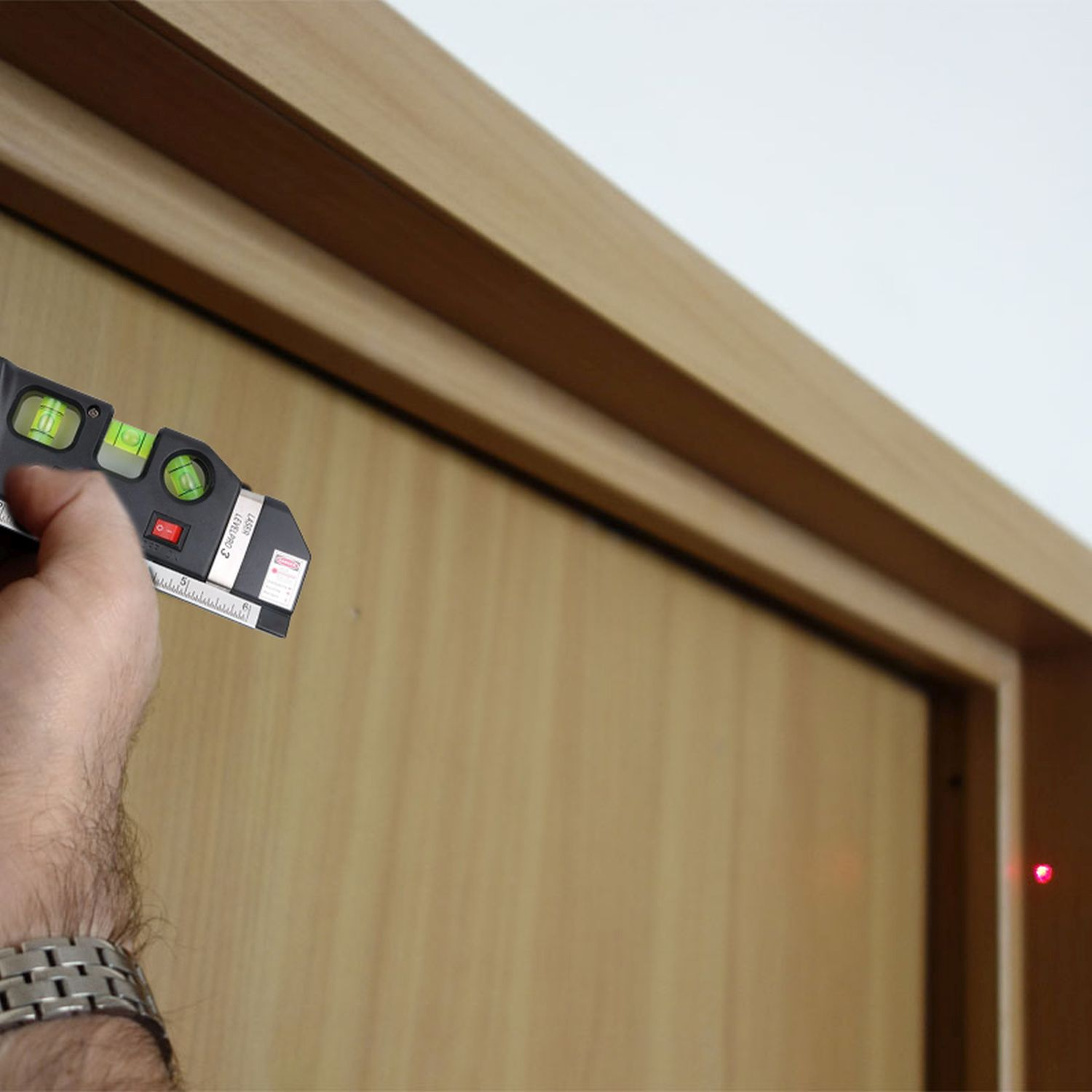 Hand hält eine Wasserwaage an einem Holzrahmen. Laserpunkt sichtbar. Weiße Wand im Hintergrund.