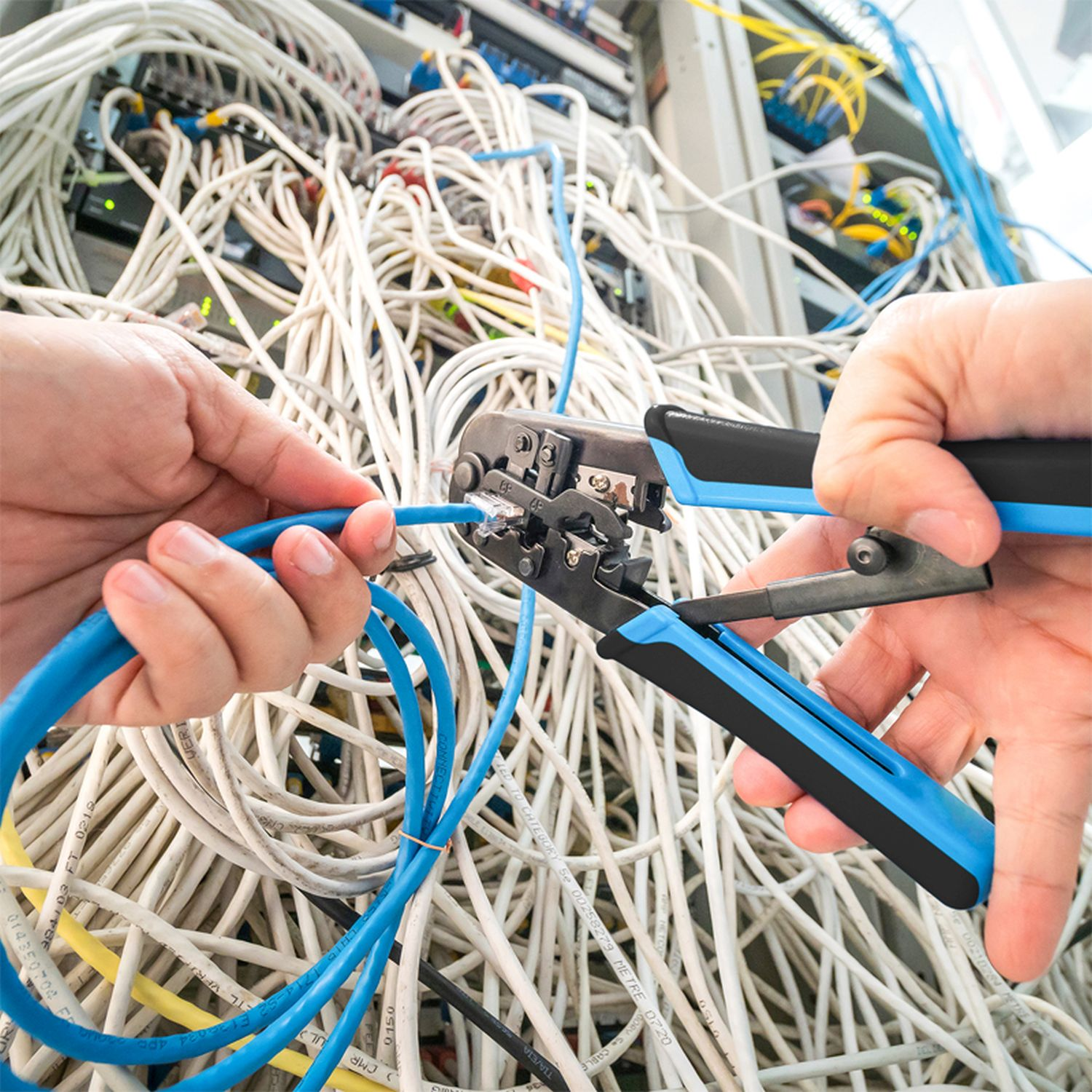 Hände, die ein blaues Netzwerkkabel mit einem Crimpwerkzeug zwischen anderen Kabeln crimpen.