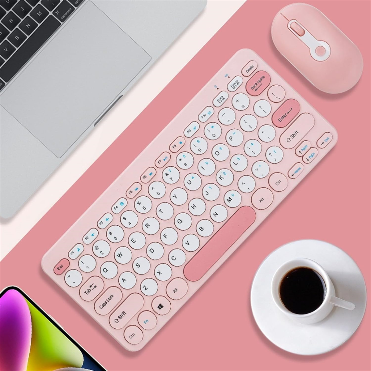 Rosa kabellose Tastatur, Maus und eine Tasse Kaffee auf rosa Oberfläche. Telefon und Laptop im Hintergrund.