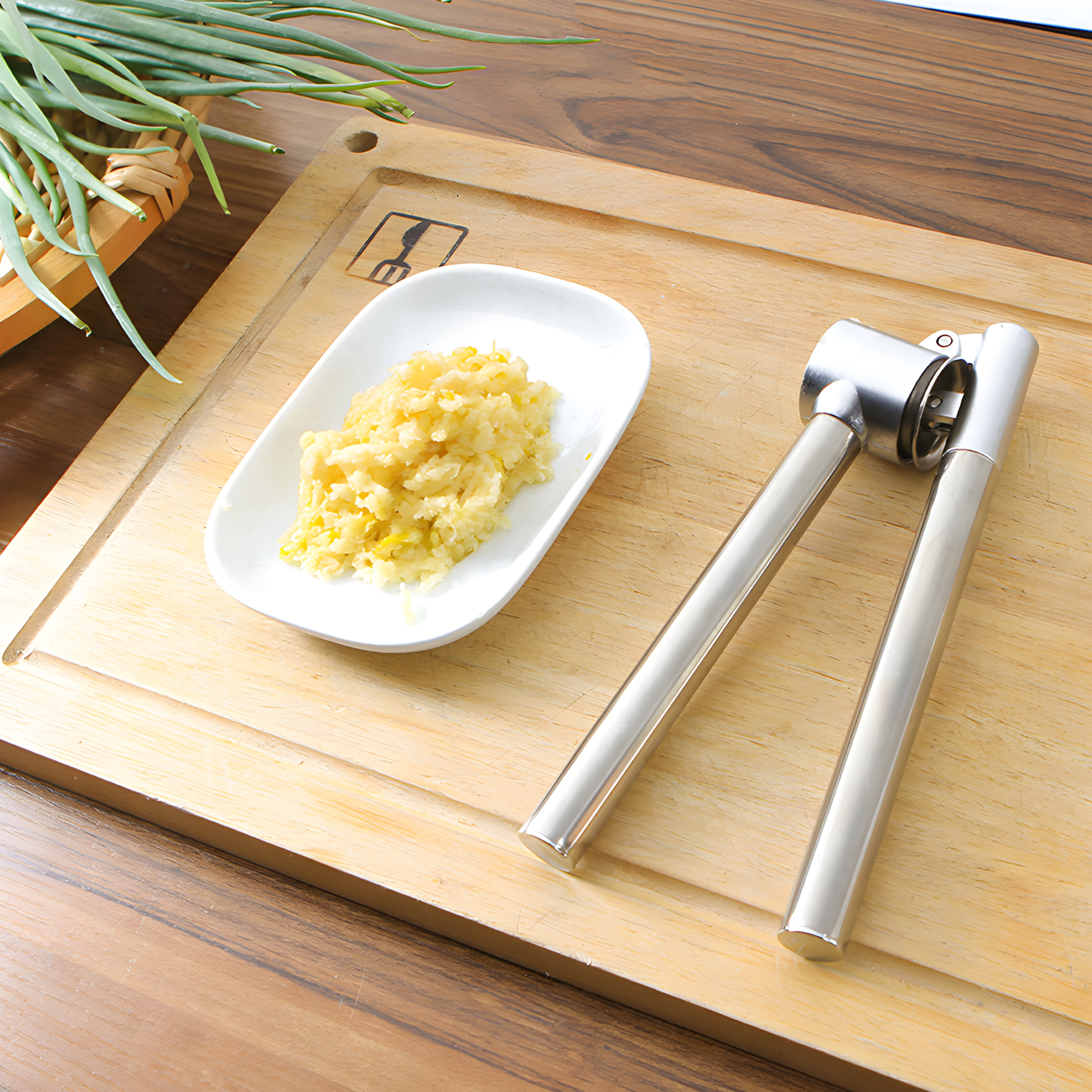 Knoblauchpresse auf Holzbrett mit gehacktem Knoblauch auf einem weißen Teller, grüne Zwiebeln hinten.