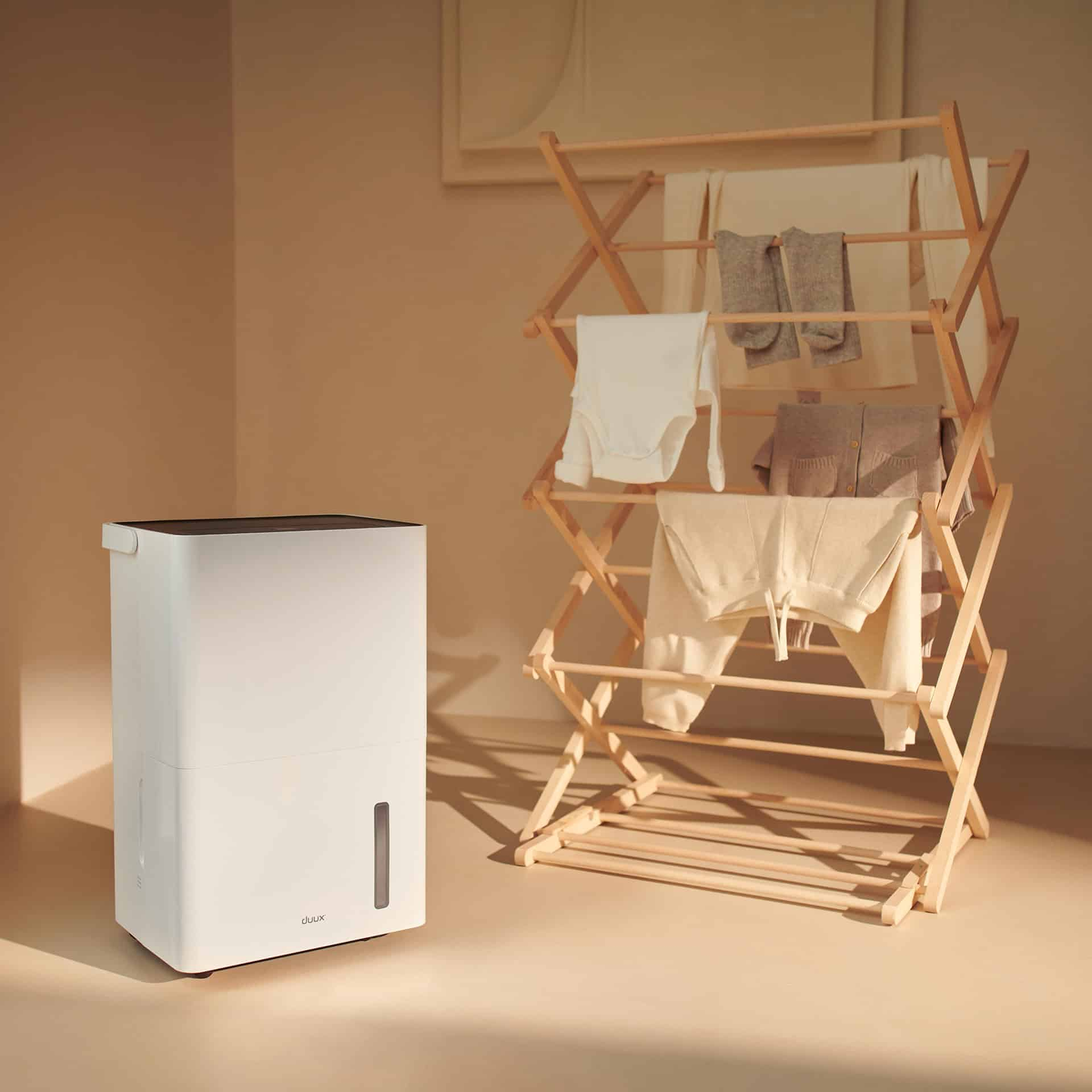 Een houten droogrek houdt opgevouwen kleding vast. Een witte luchtreiniger staat op de vloer.