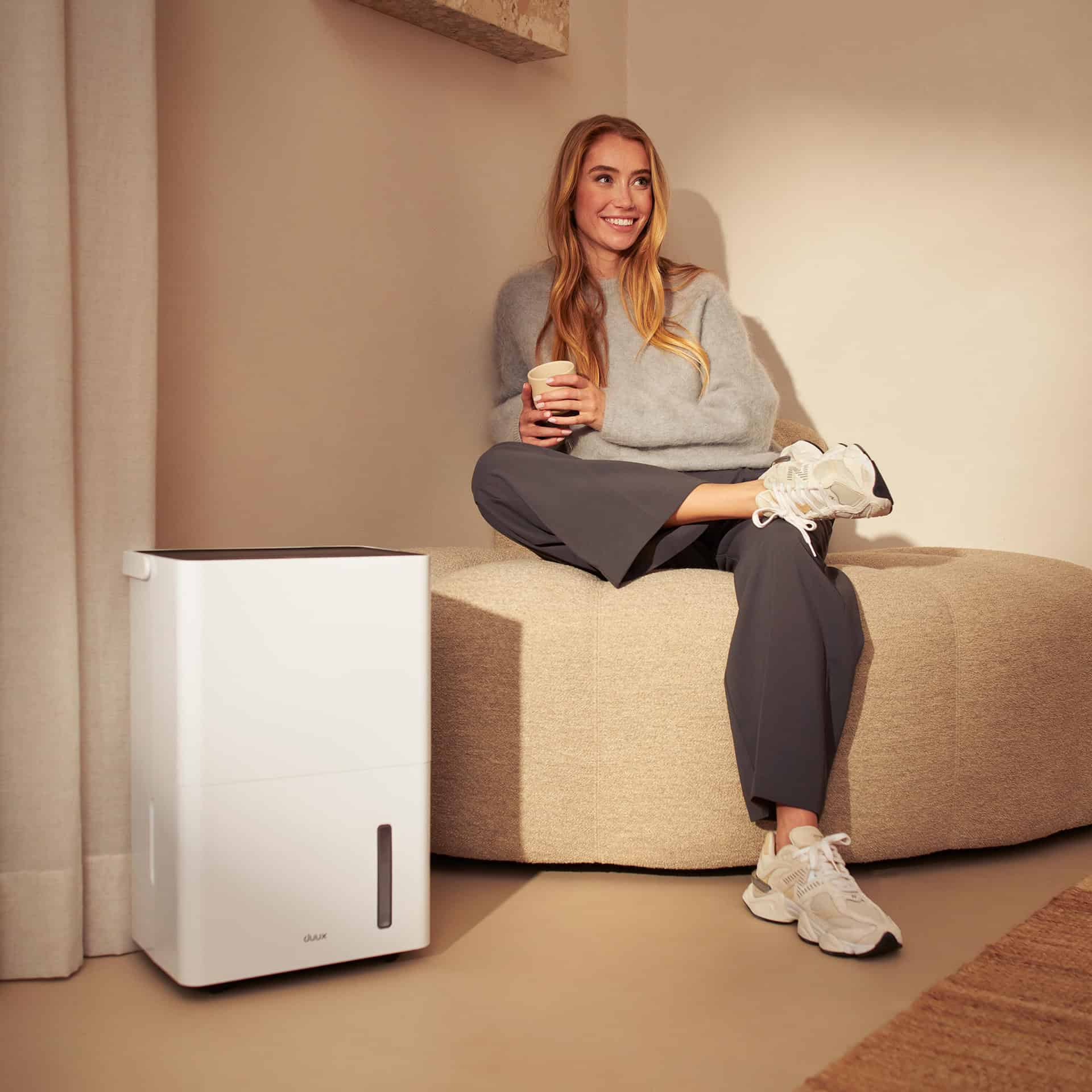 Vrouw zittend met een luchtontvochtiger. Ze houdt een kopje vast, glimlacht. Beige en witte objecten.