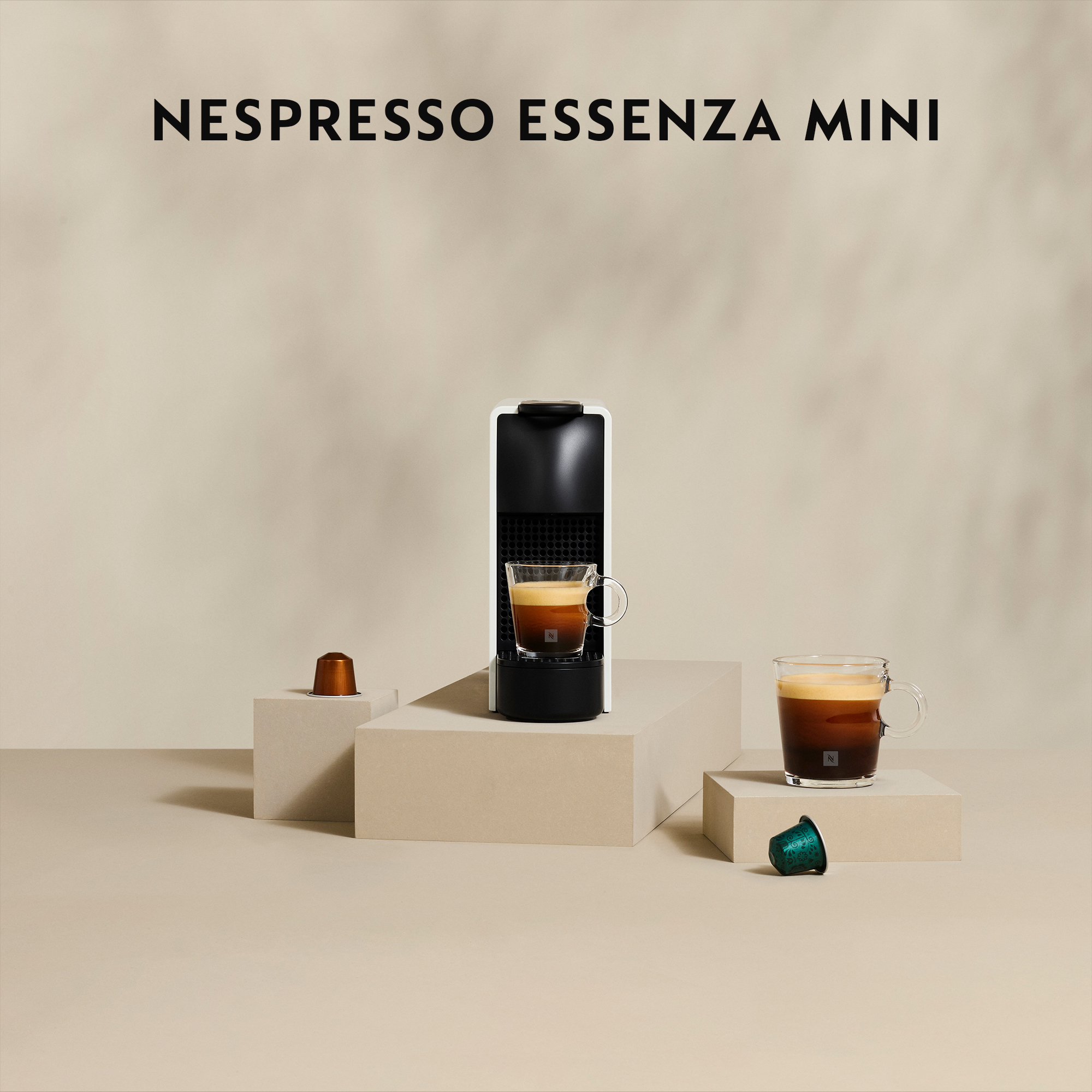Ekspres do kawy Nespresso Essenza Mini z filiżanką kawy na jasnym tle. Widoczne są dwie kapsułki.