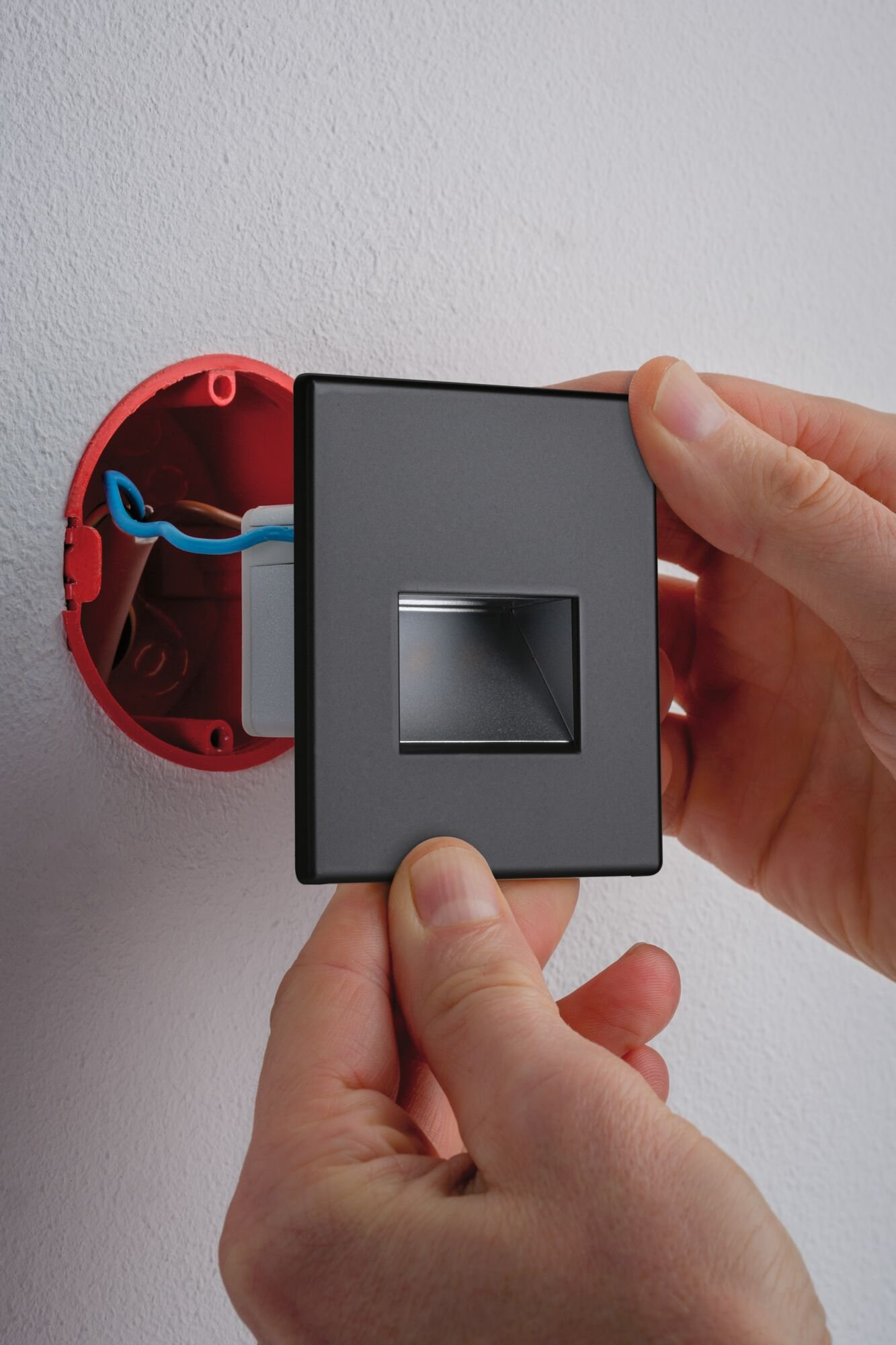 Les mains installent une prise électrique gris foncé dans une boîte électrique rouge sur un mur blanc.