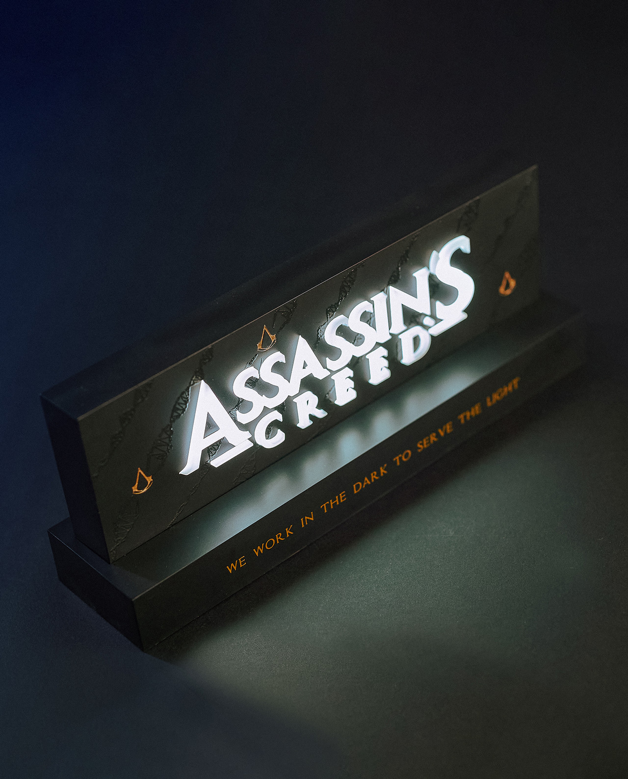 Beleuchtetes Schild 'ASSASSINS CREED'. Schwarze Basis mit goldenen Akzenten. Dunkler Hintergrund. Text: 'WE WORK...'.