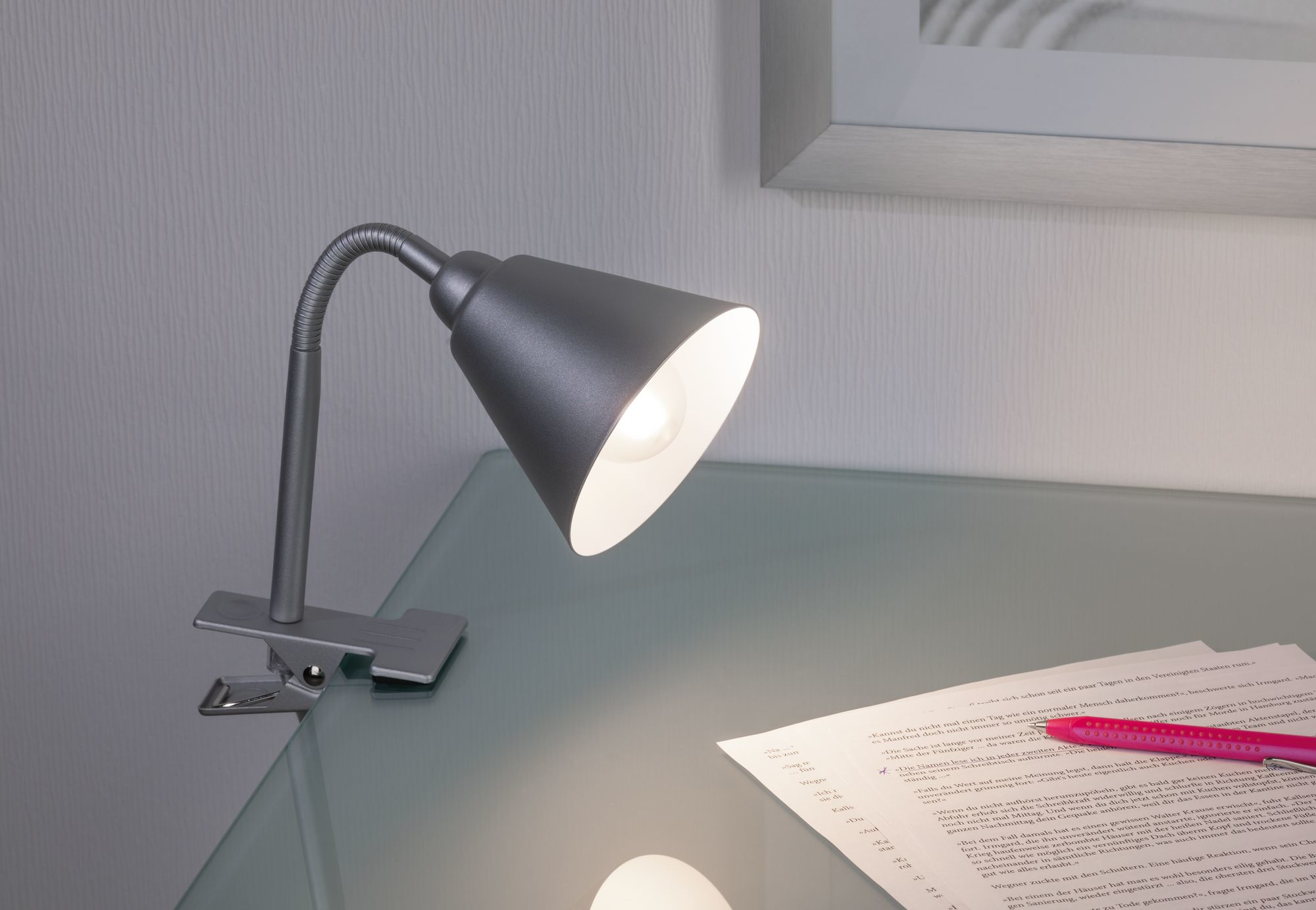 Lampa biurkowa z elastyczną szyją jest przymocowana do szklanego stołu. Na stole są papiery i długopis.