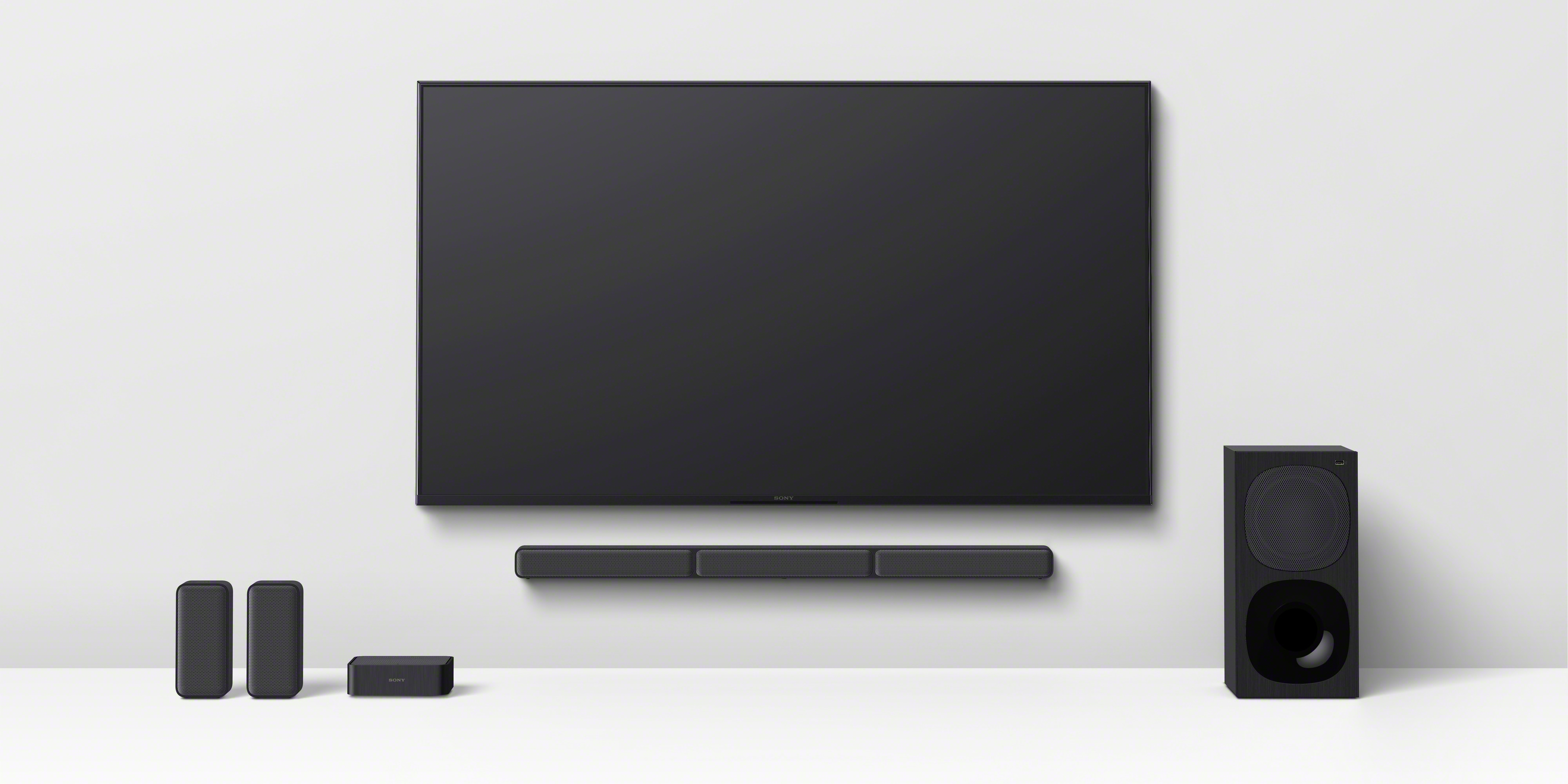 Fernseher an der Wand. Soundbar darunter. Lautsprecher an den Seiten. Schwarzer Subwoofer rechts.