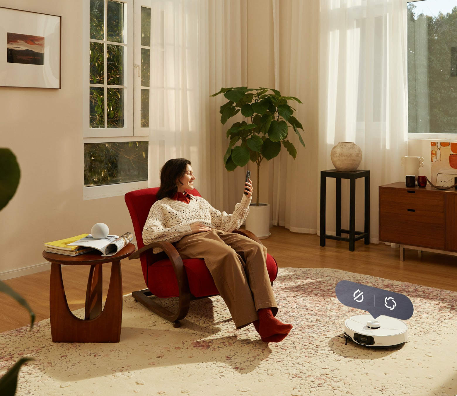 Kobieta siedzi na krześle, używając telefonu. Odkurzacz robot jest na podłodze. Okno jest w tle.