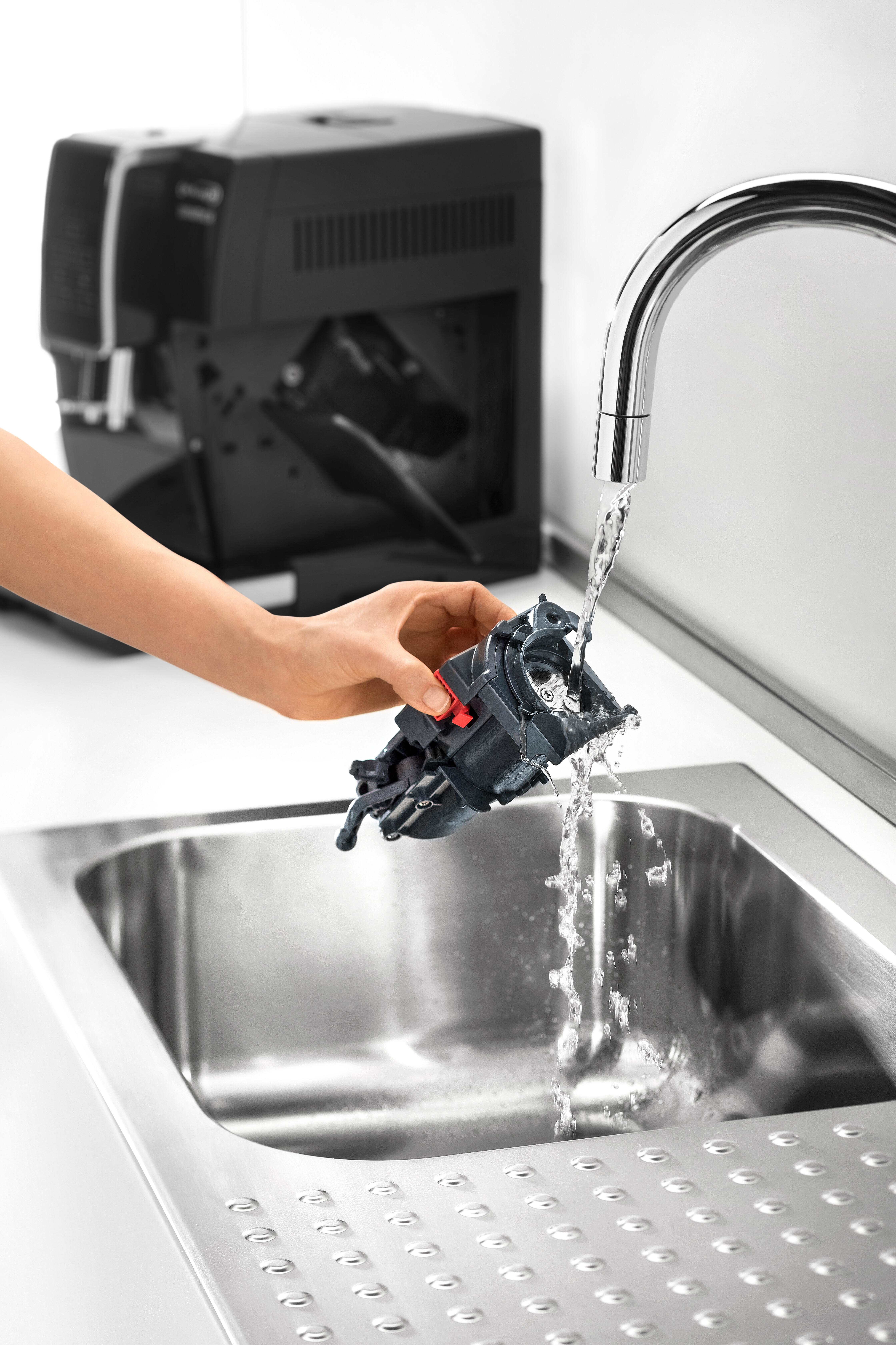 Eine Hand wäscht ein Kaffeemaschinenteil unter einem Wasserhahn aus Metallbecken.