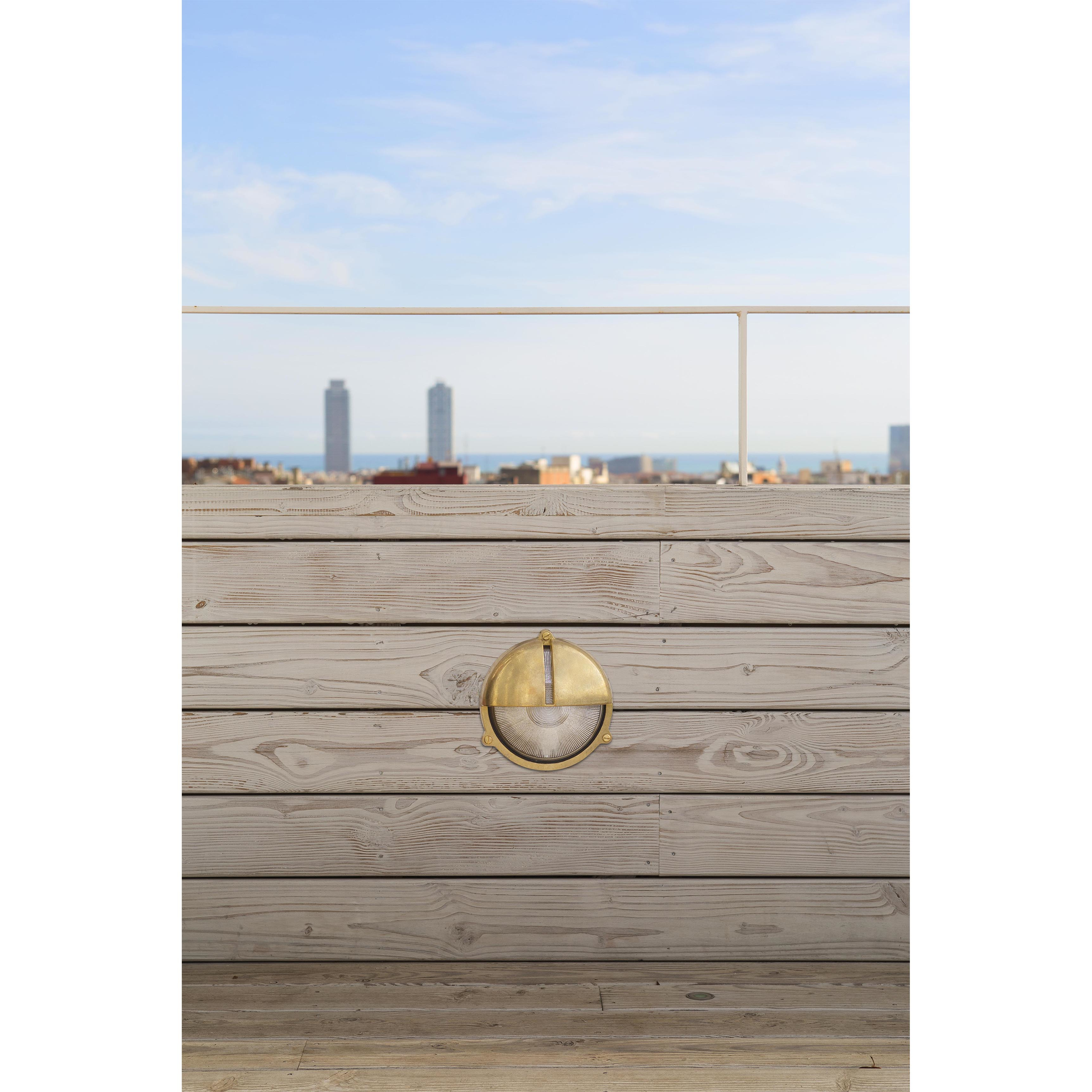 Un apparecchio di illuminazione rotondo in ottone su una parete di legno. Uno skyline e cielo blu sullo sfondo.