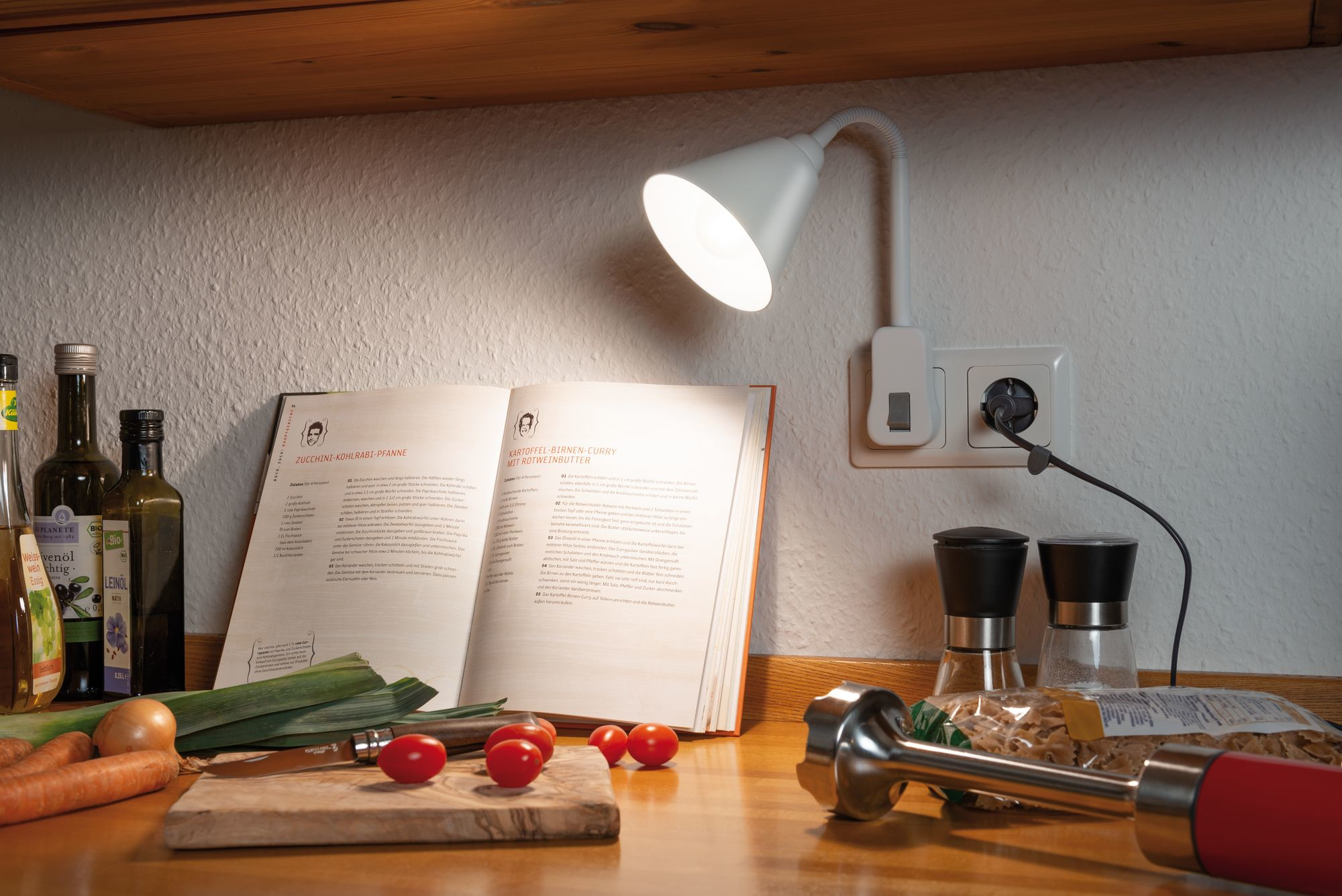 Scena della cucina con una lampada, un libro di cucina aperto, verdure e un frullatore rosso.