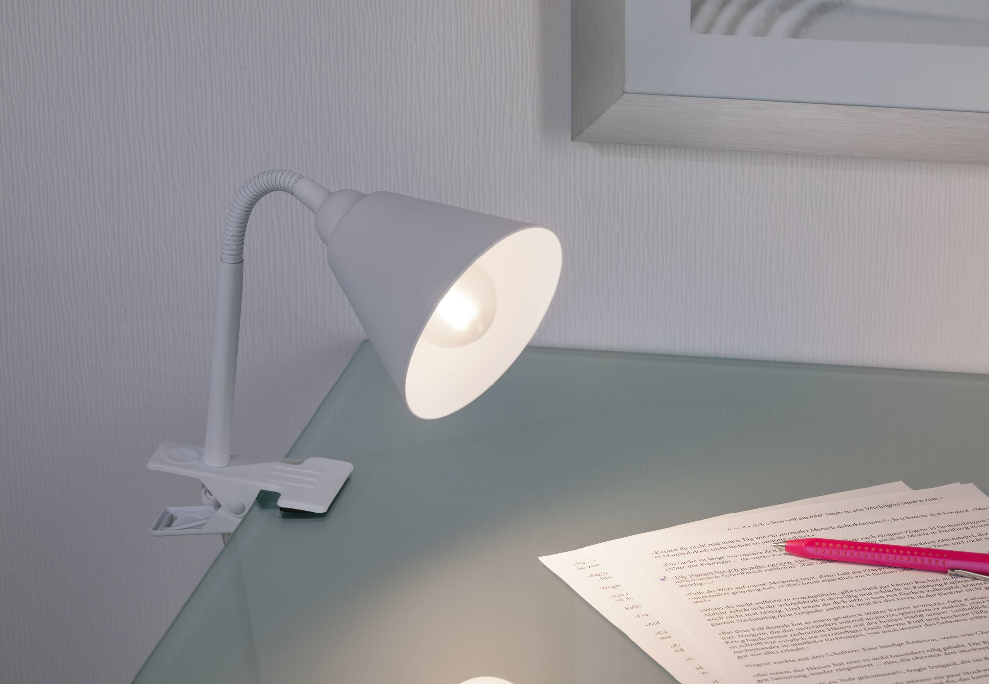 Eine weiße Schreibtischlampe mit flexiblem Hals, die Licht auf Papier auf einer Glasoberfläche scheinen lässt. Es gibt einen rosa Stift.