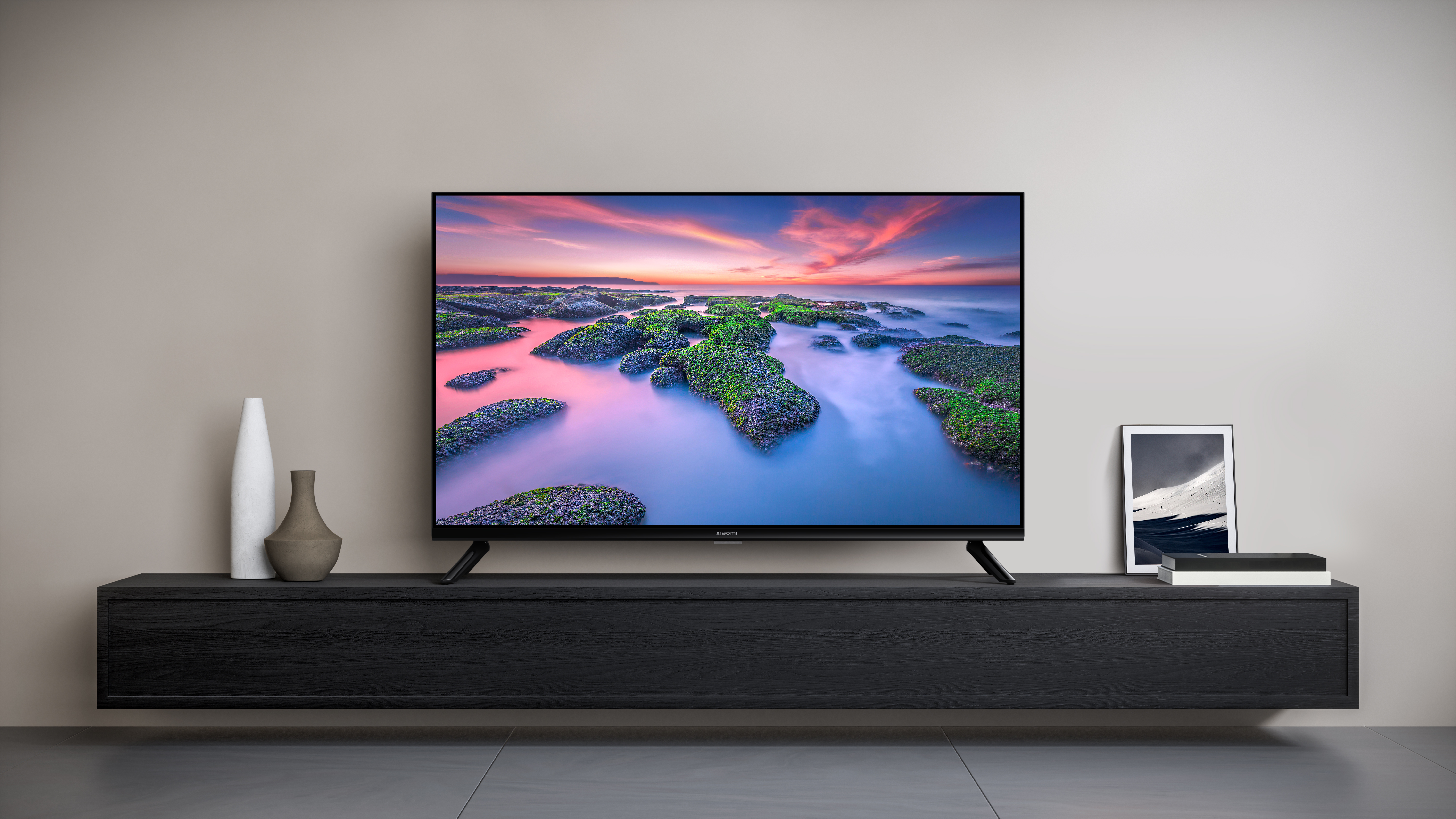 Telewizor wyświetlający scenę plażową, ustawiony na czarnym stojaku na tle beżowej ściany.