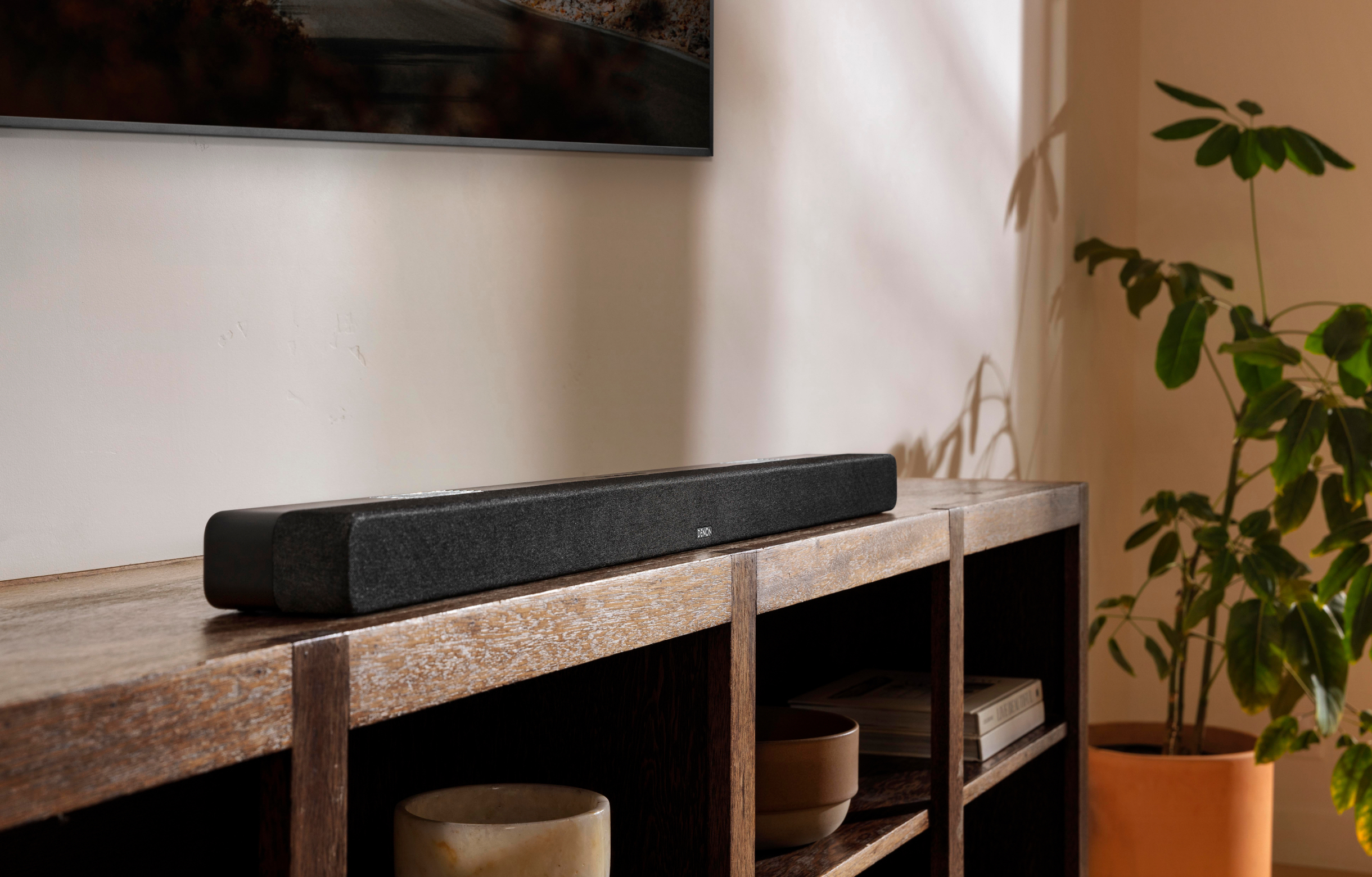 Schwarze Soundbar auf Holzschrank, neben Pflanze. TV im Hintergrund.