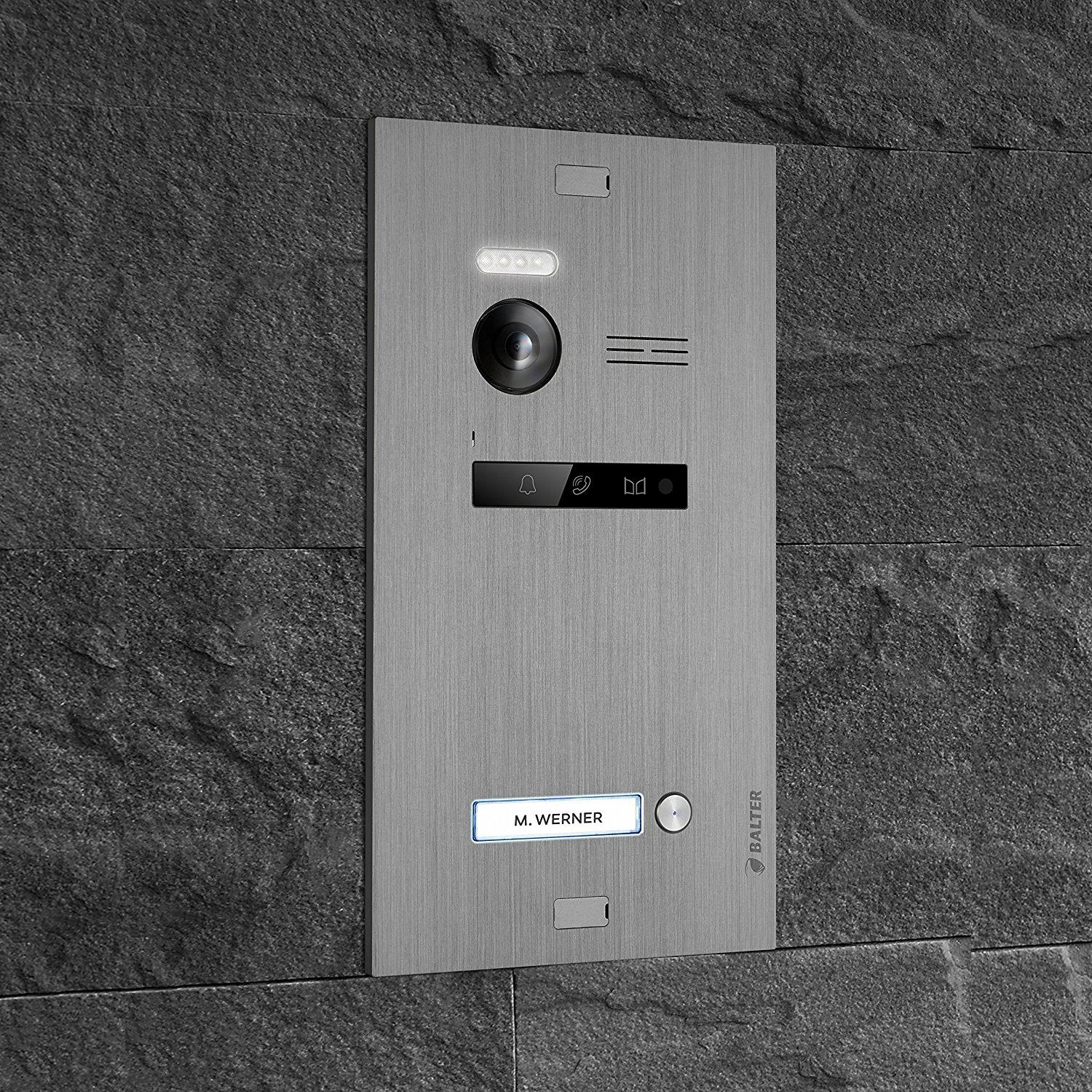 Ein silbernes Türsprechanlagensystem an einer dunklen Steinmauer. Enthält eine Kamera, einen Lautsprecher und ein Namensschild.