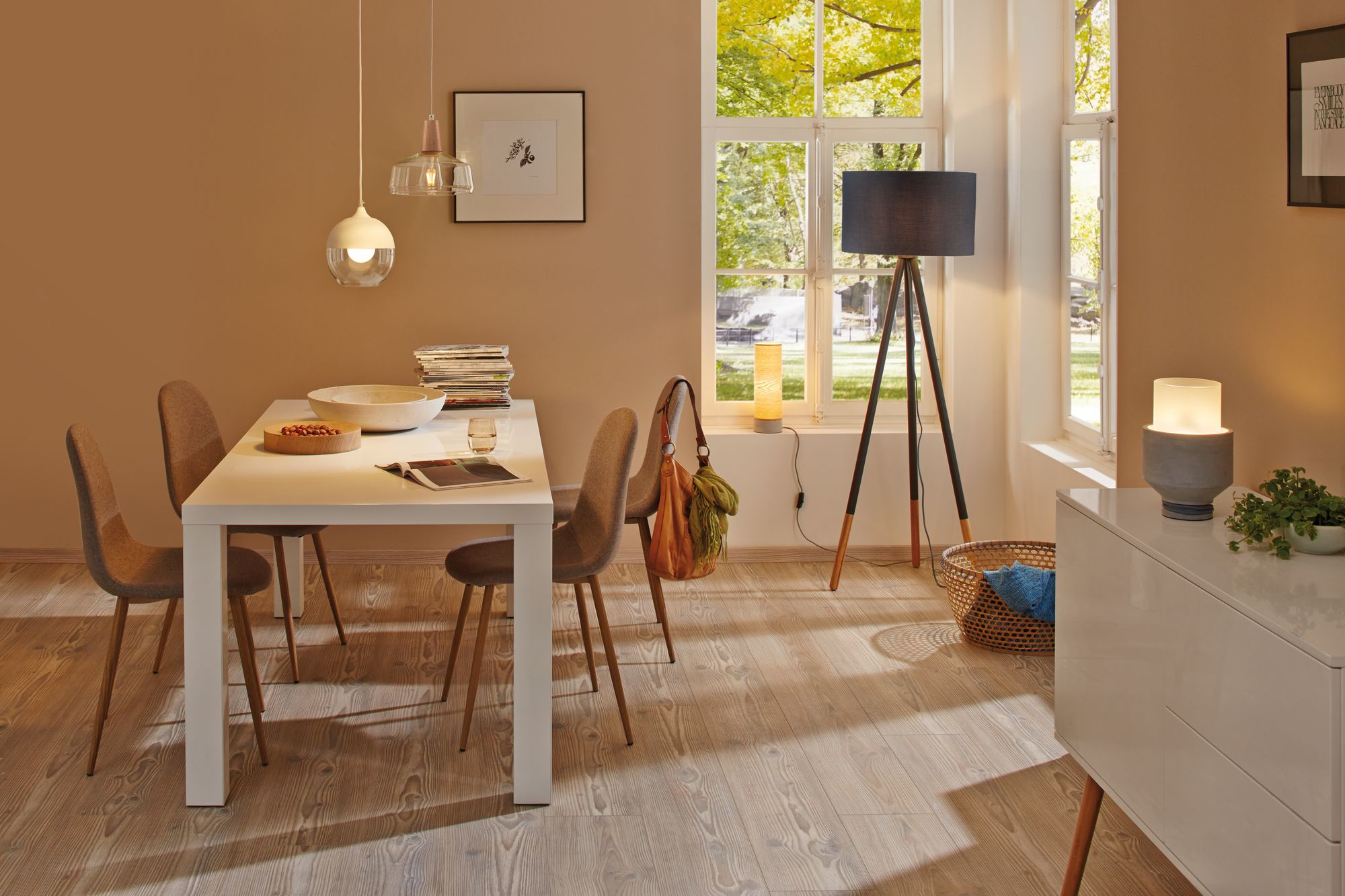 Esszimmer: weißer Tisch, Stühle, Lampen, Fenster und Sideboard. Warmes Farbschema.