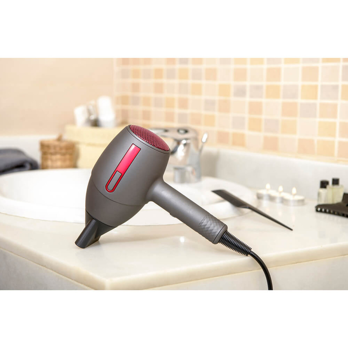 Un secador de pelo gris y rojo está sobre una superficie blanca en un baño, cerca de un lavabo y algunos artículos de tocador.