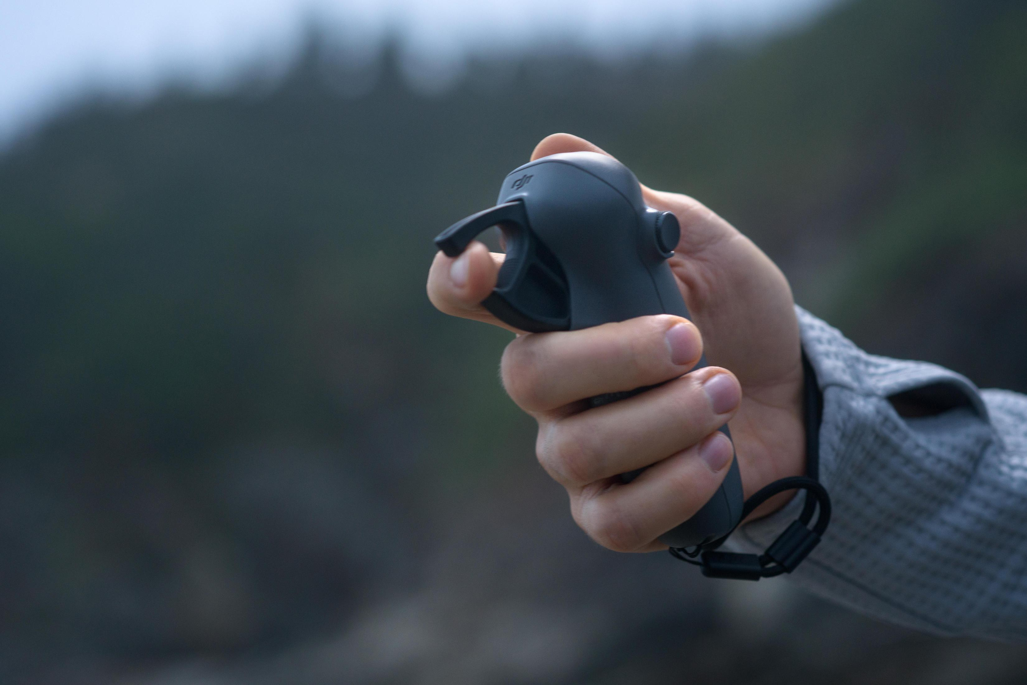 Dłoń trzymająca czarny kontroler drona. Tło jest rozmyte z zielonymi i szarymi tonami.