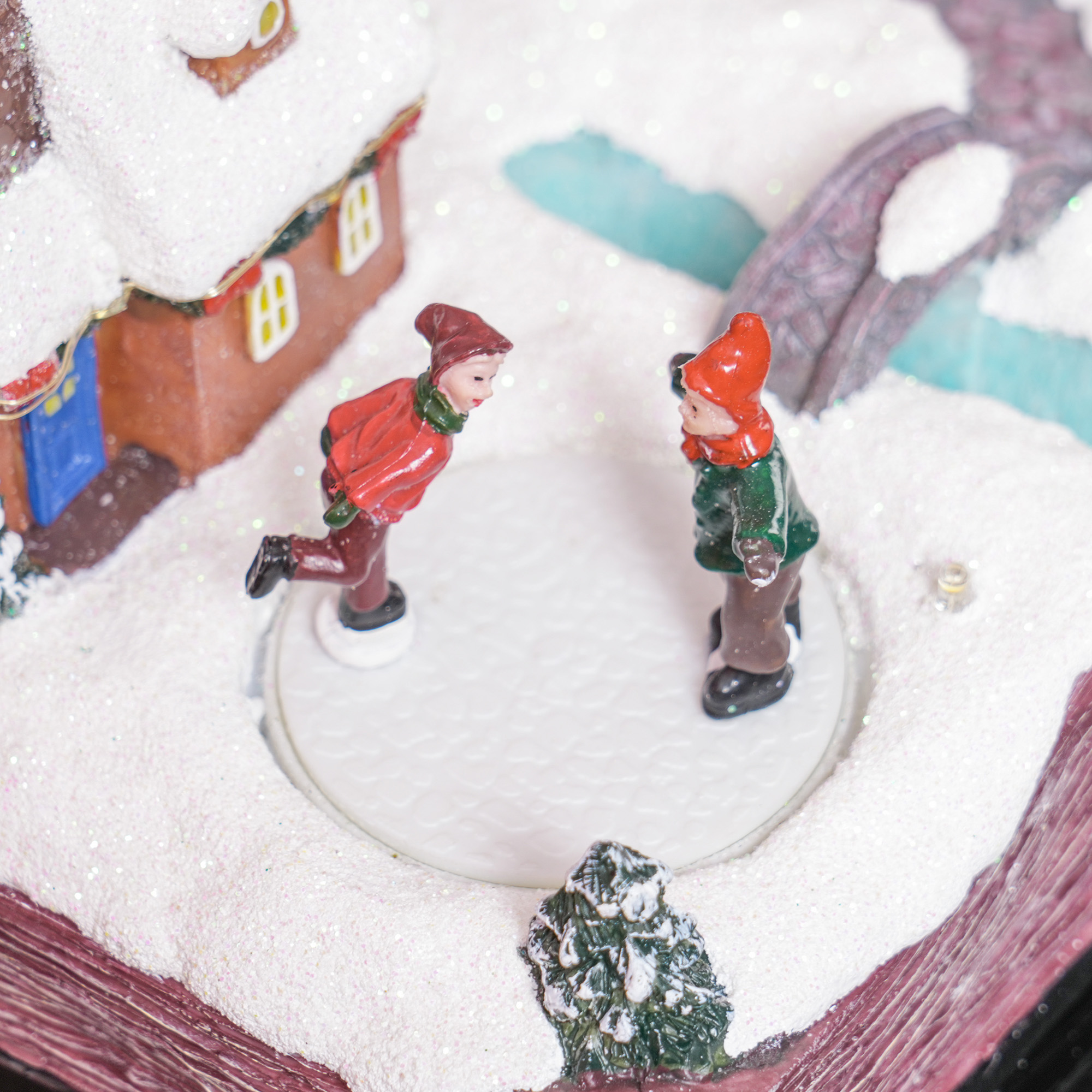 Dos pequeñas figuras patinando sobre hielo en una superficie blanca rodeada de nieve, dentro de una decoración navideña.