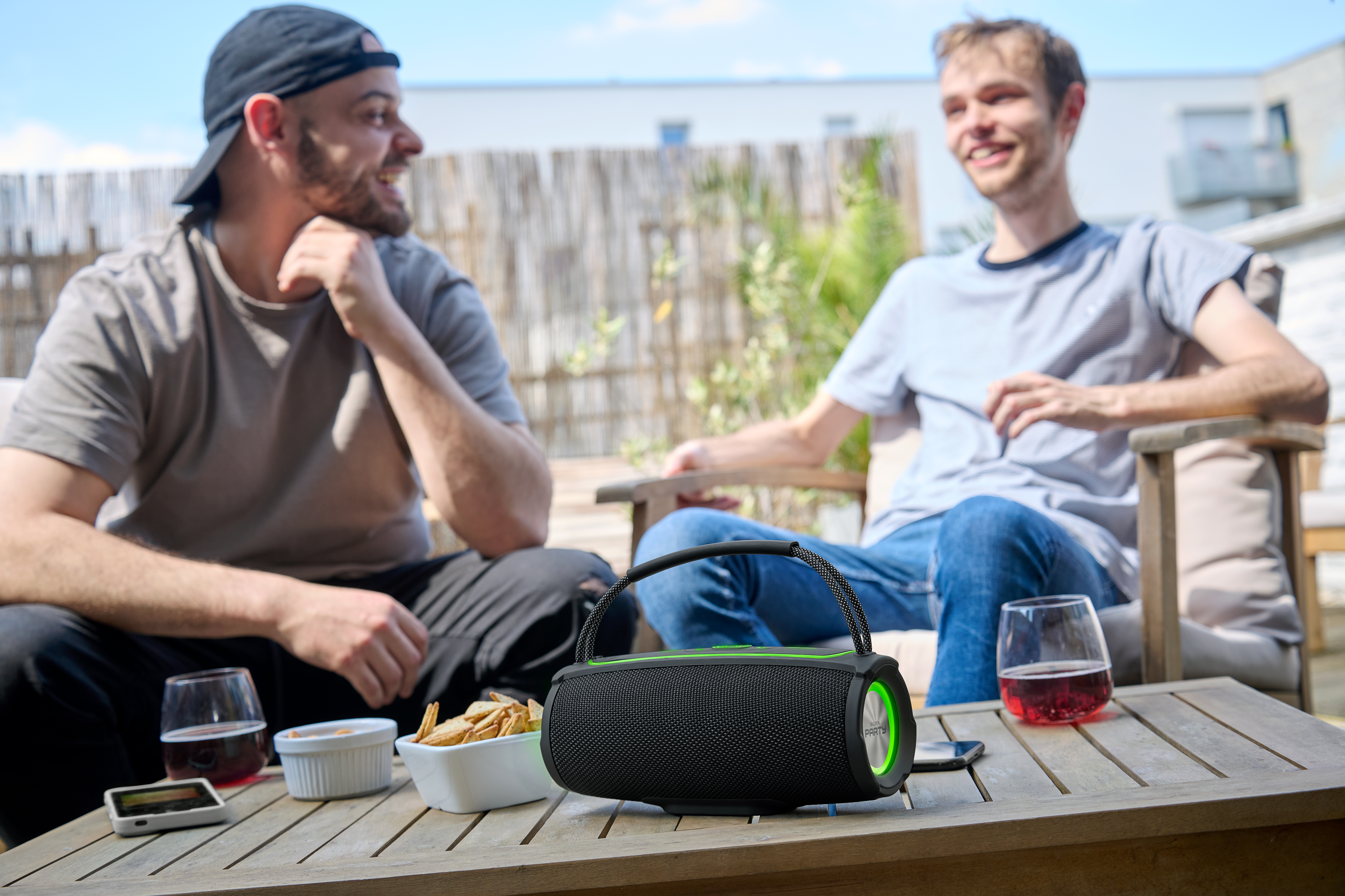 Zwei Männer sitzen draußen und lächeln. Ein tragbarer Lautsprecher und Getränke stehen auf einem Tisch vor ihnen.