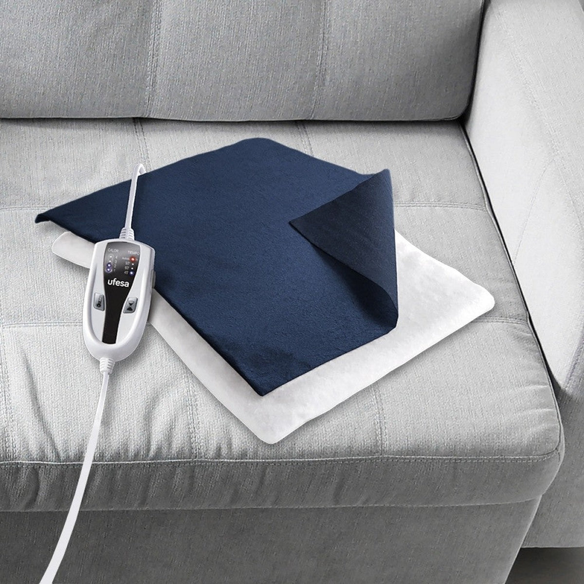 Niebiesko-biała poduszka grzewcza na szarej kanapie. Posiada panel sterowania i kabel.