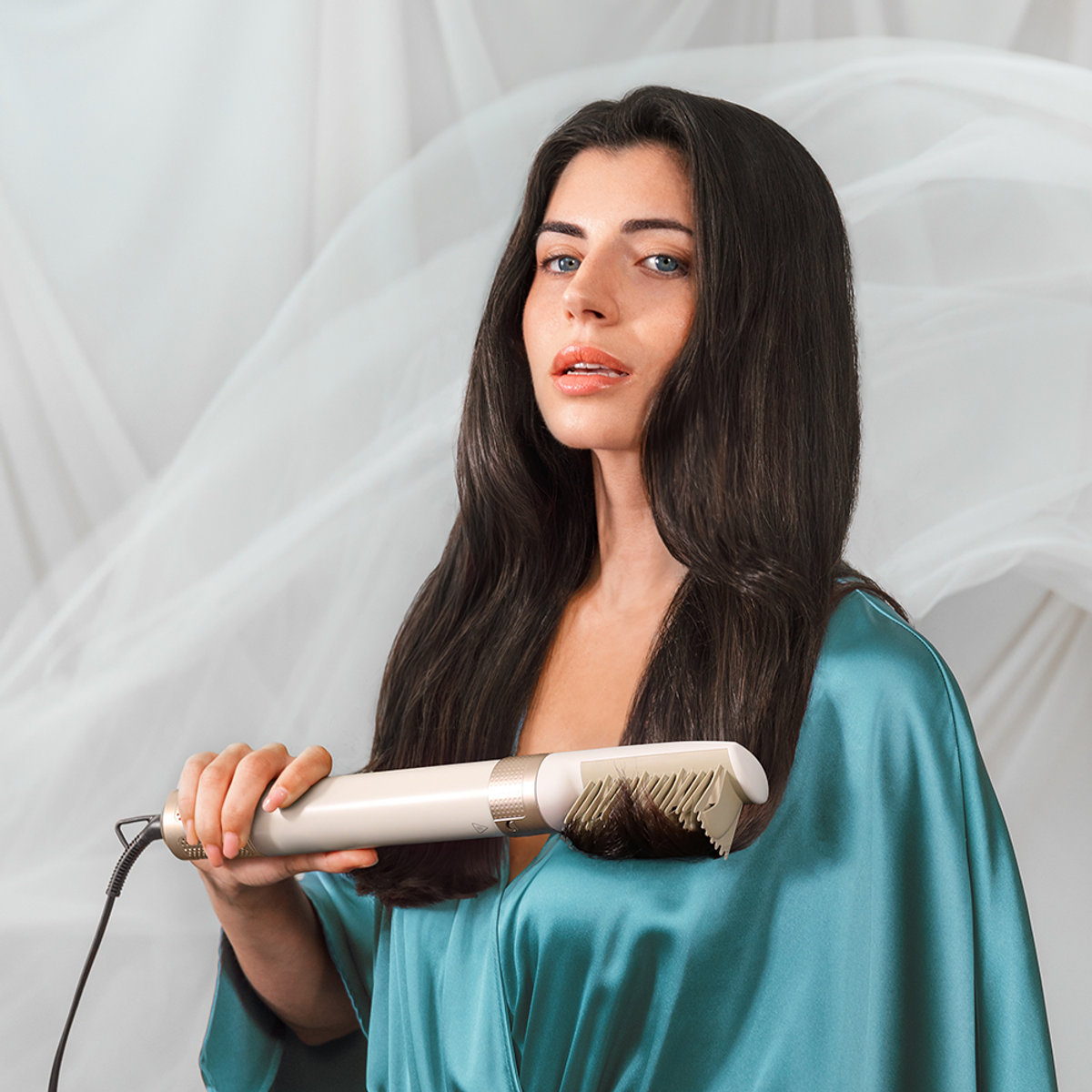 Donna che tiene uno strumento per lo styling dei capelli. Indossa un accappatoio turchese davanti a uno sfondo bianco sfocato.