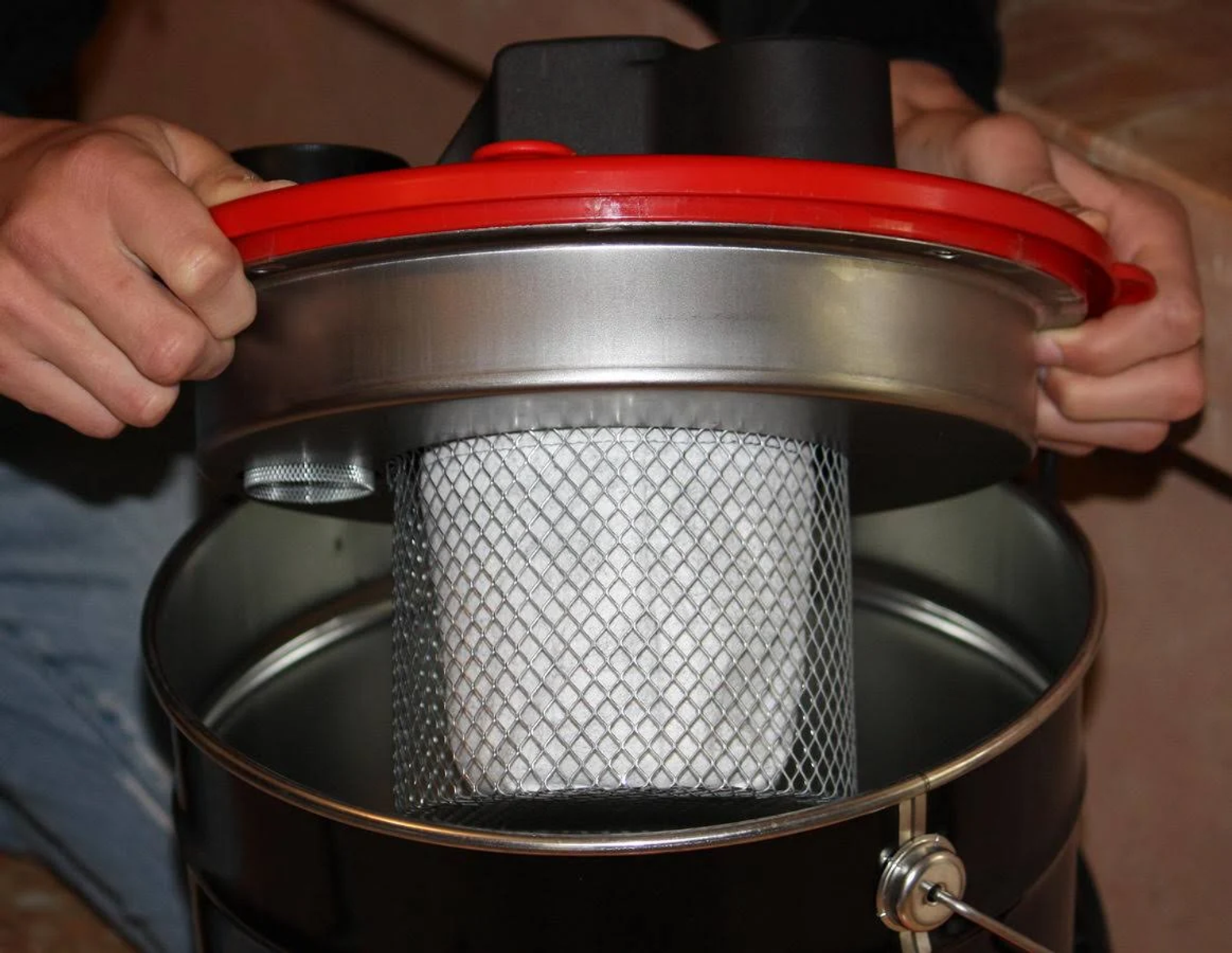 Ręce trzymające filtr nad czarnym odkurzaczem. Filtr jest biały i metalowy.