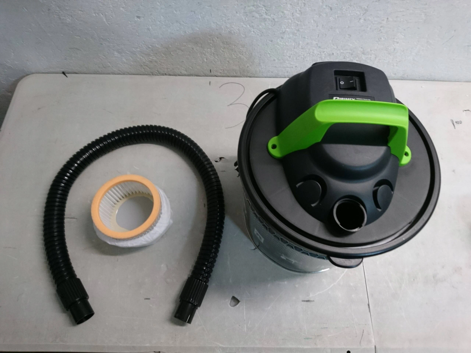 Un aspirapolvere umido/secco con tubo, filtro e maniglia verde. Componenti su superficie chiara.