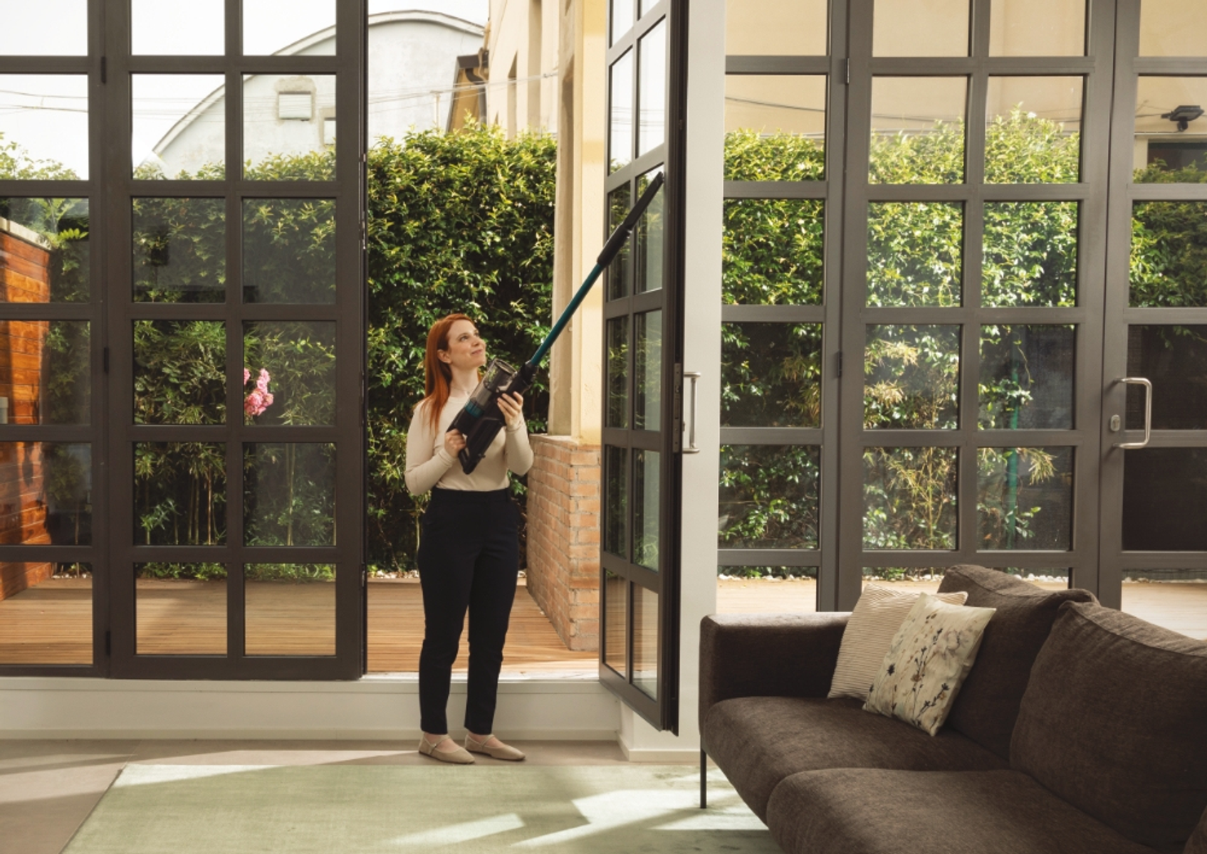 Una mujer limpiando una ventana con una aspiradora, interior con un sofá.