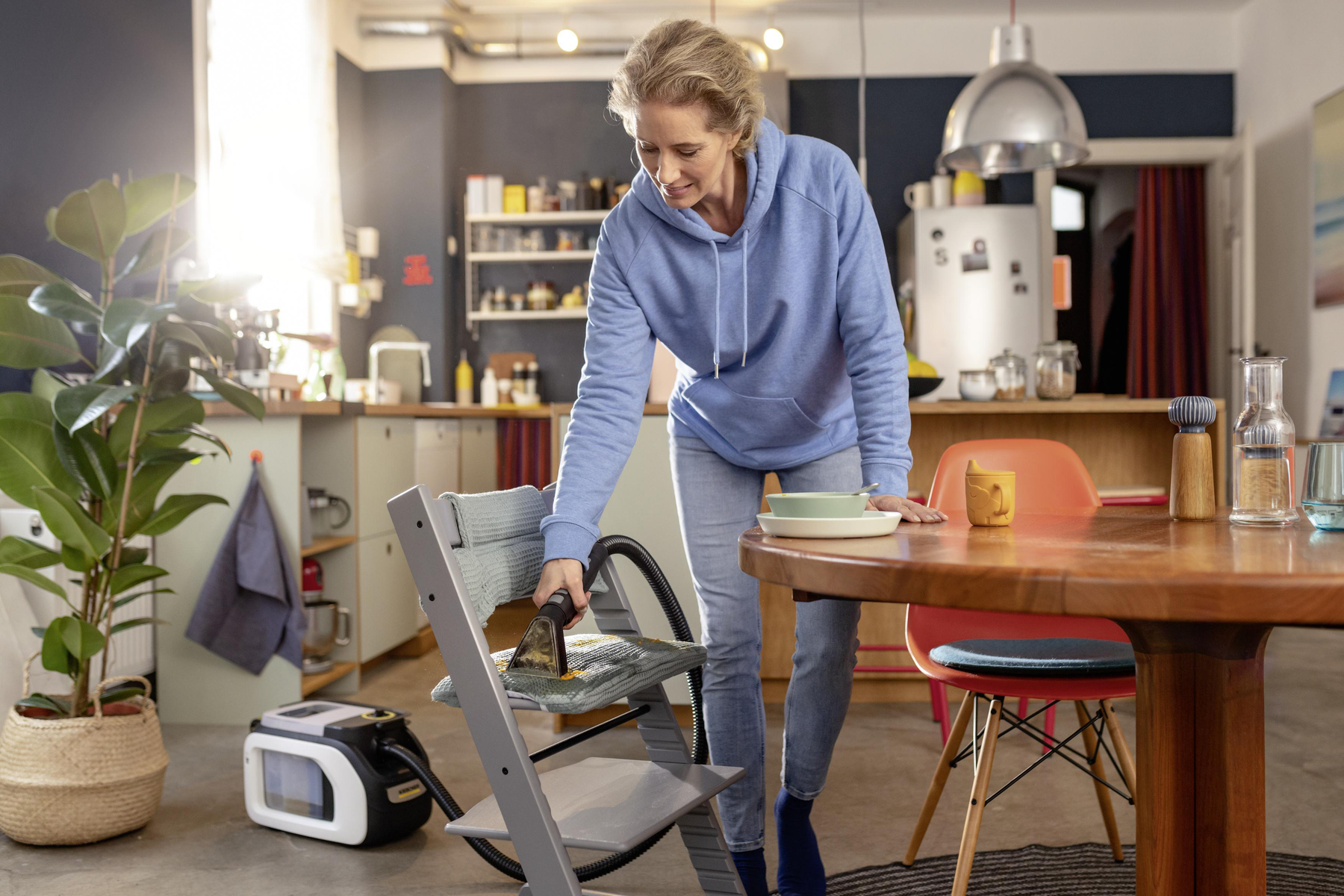 Kobieta czyszcząca krzesełko do karmienia odkurzaczem w kuchni.