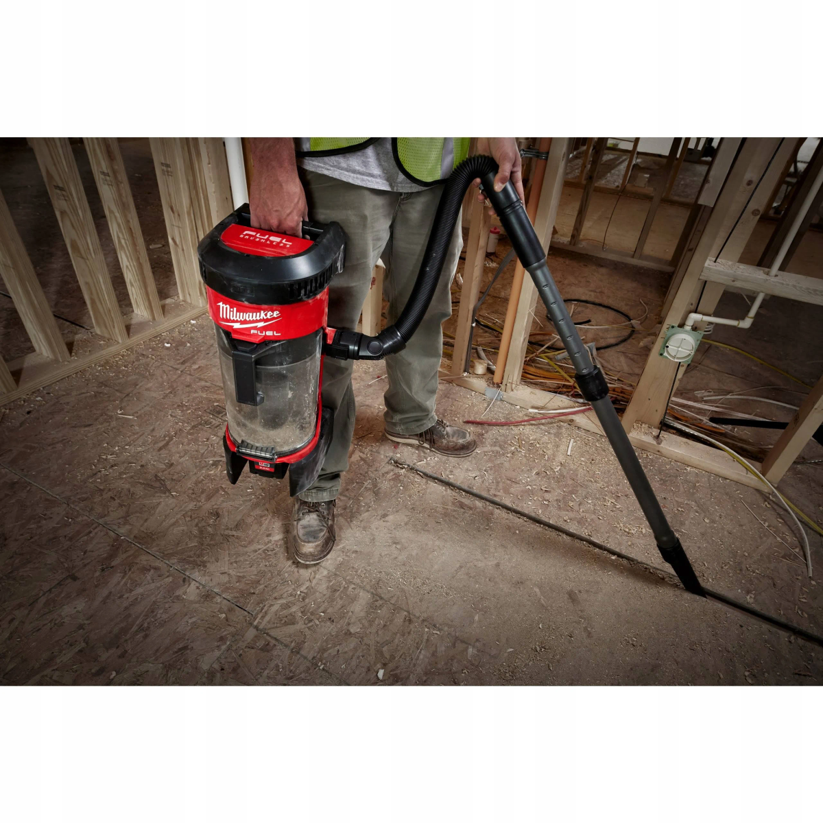 Un uomo tiene un aspirapolvere rosso e nero. La scena è un cantiere con struttura in legno.