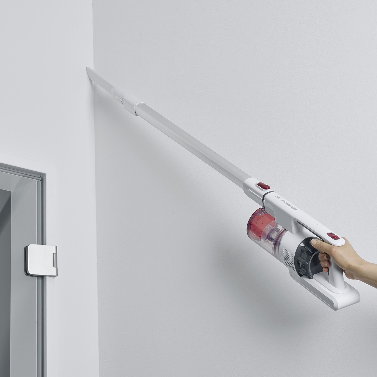 Un aspirapolvere bianco senza fili pulisce un muro bianco. Accenti rossi. Mano visibile. Stipite della porta a sinistra.
