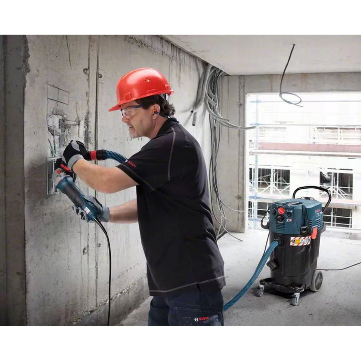 Un trabajador con casco usando una herramienta en una pared. Una aspiradora está conectada a la herramienta.