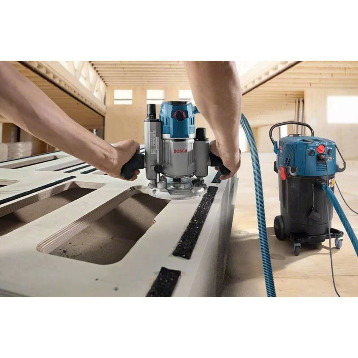 Persona usando una fresadora azul sobre madera, con una aspiradora cerca.