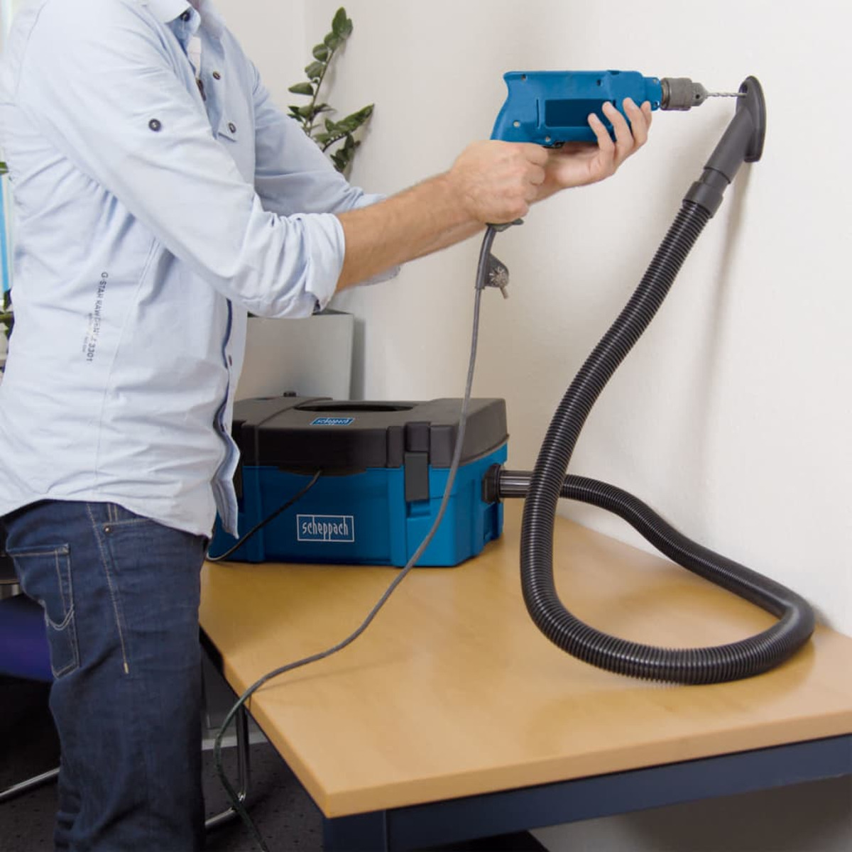 A man drilling into a wall with a blue drill. A blue vacuum sits on a table.