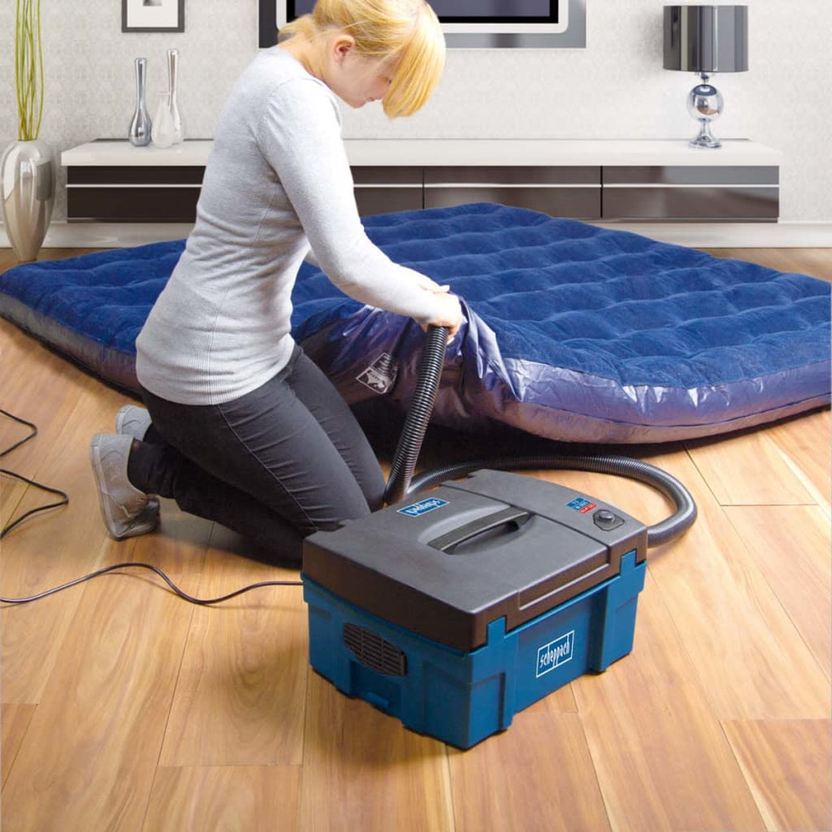 Woman inflating a blue air mattress with an electric pump on a wooden floor.