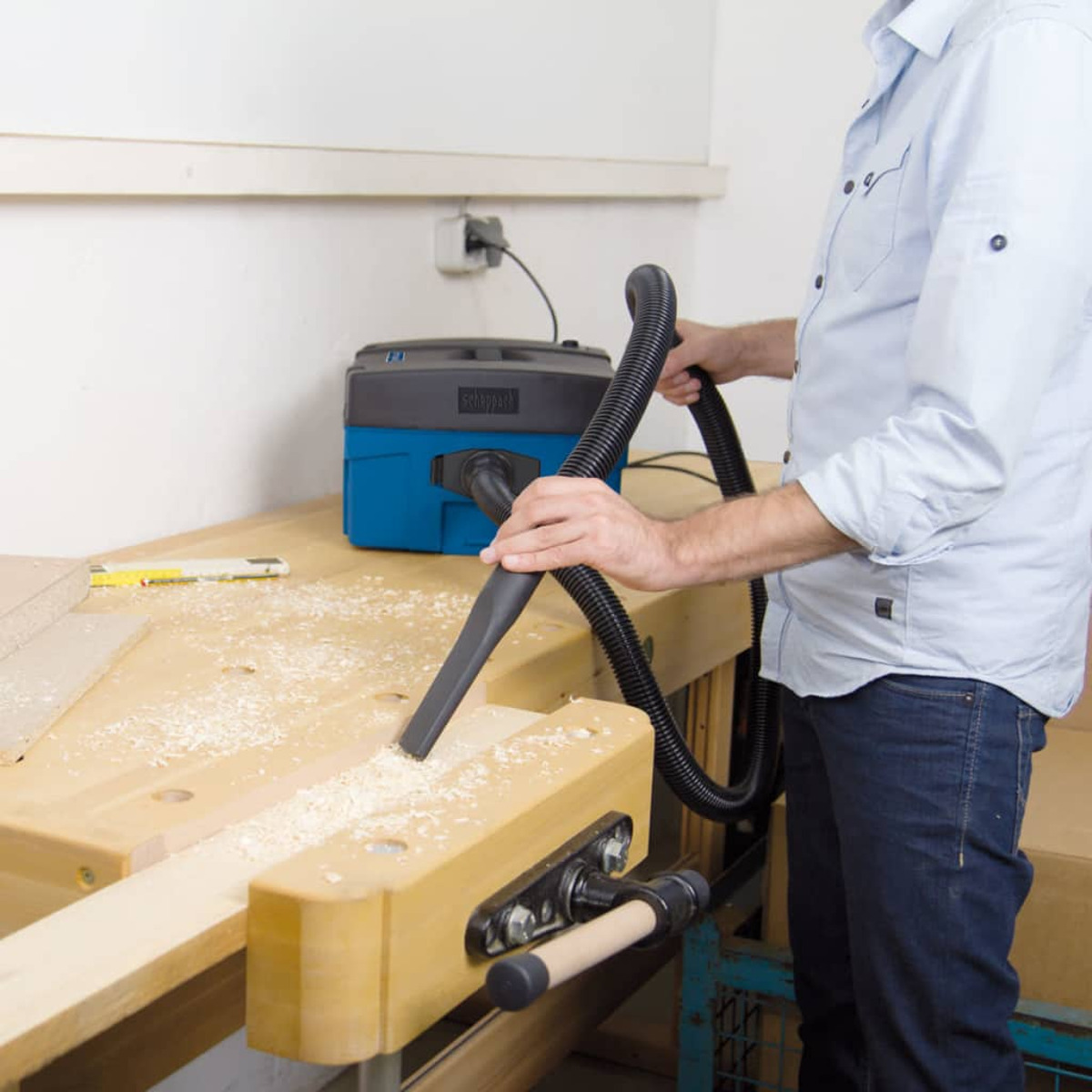 Man using a vacuum cleaner on a wooden workbench, wood shavings visible.