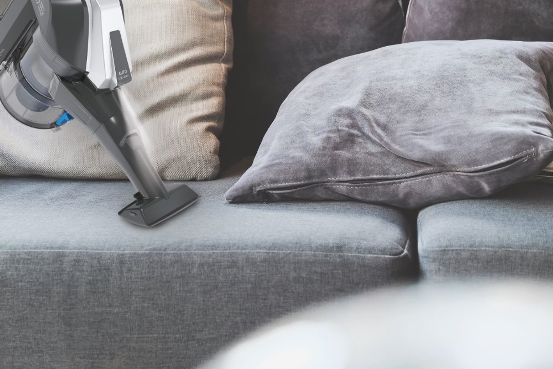 Una persona che aspira un divano grigio con un aspirapolvere portatile. La testina è visibile.