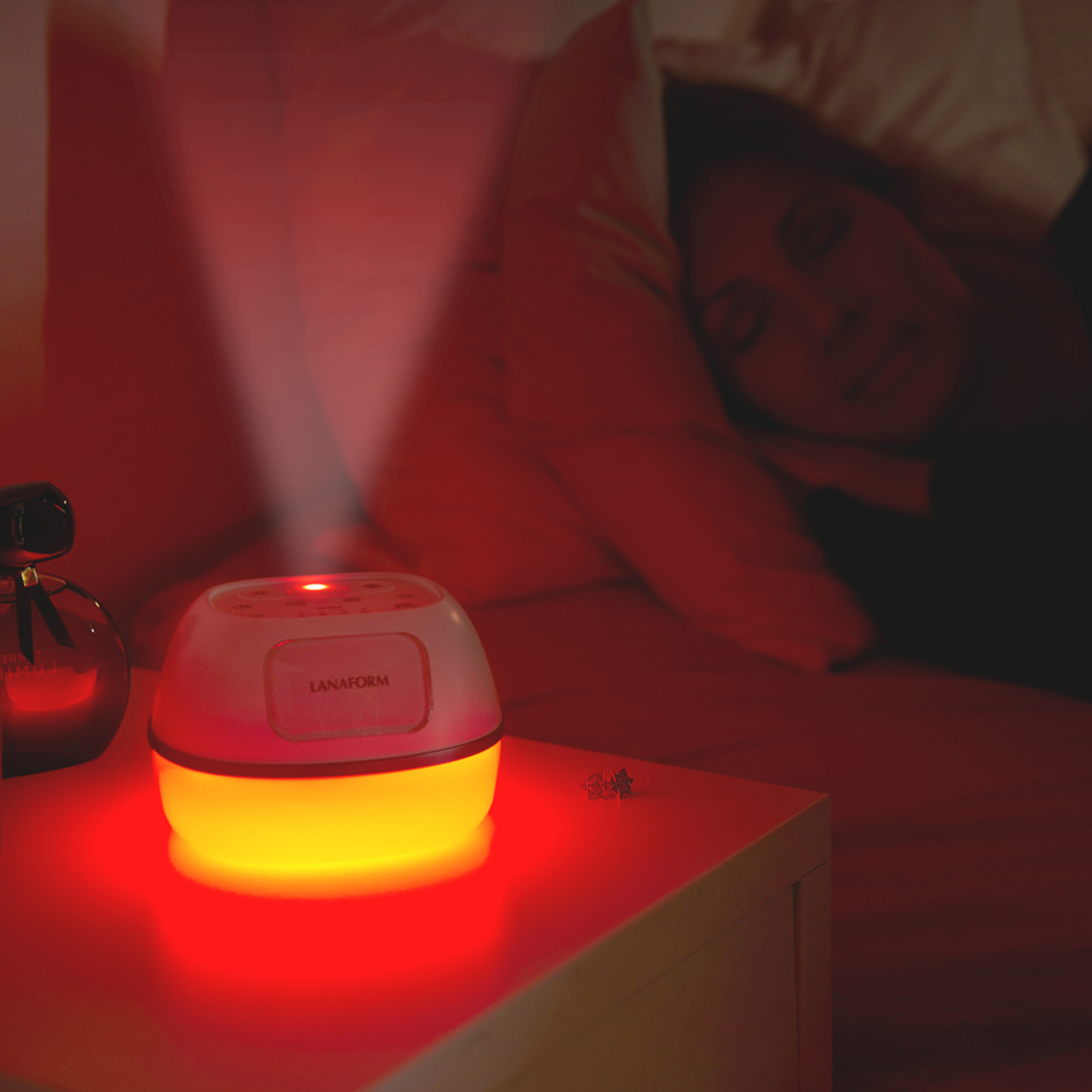 A white and orange lamp projects a red light into a dark room. A person is sleeping in the background.