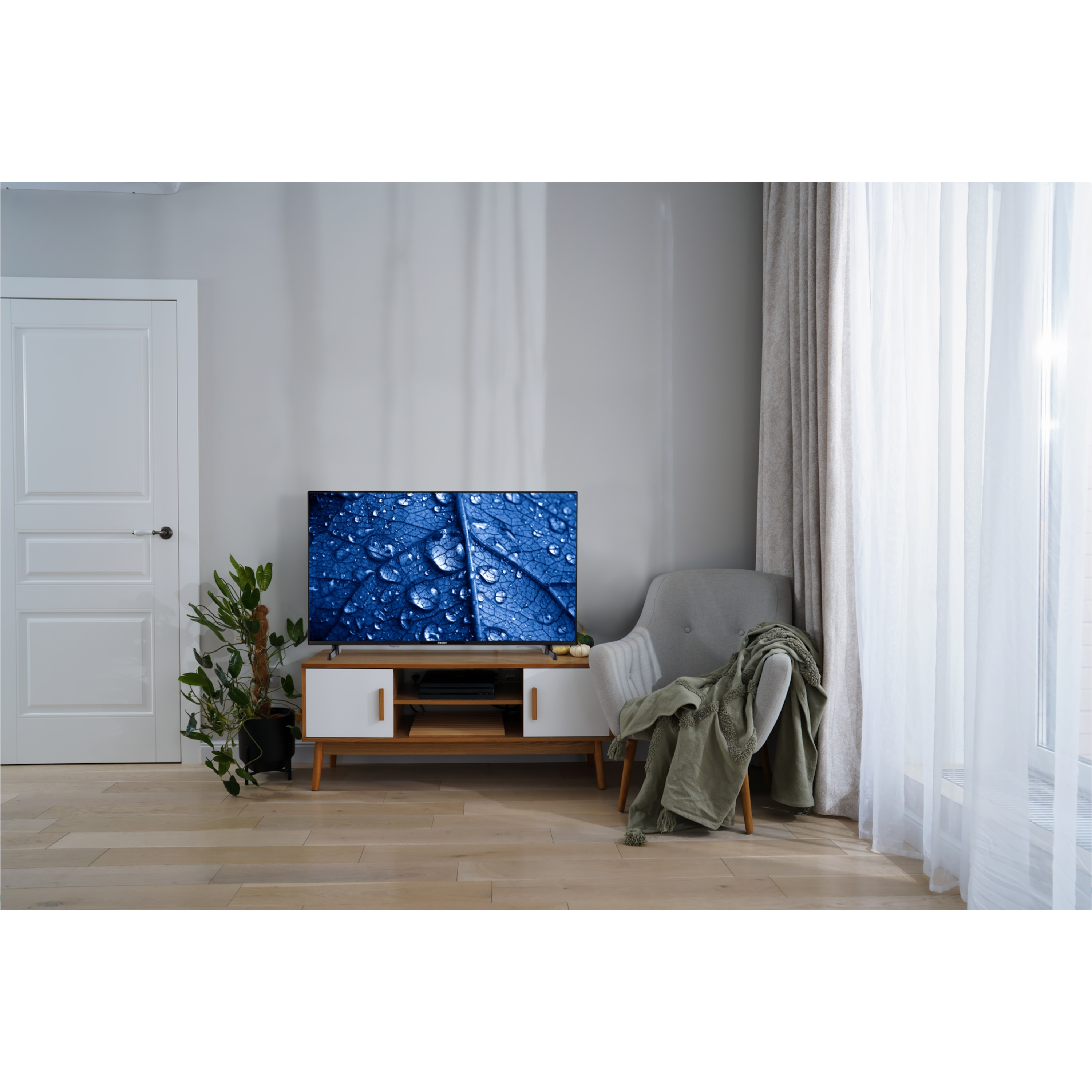 Een tv op een witte kast, een plant, een grijze stoel en een raam met gordijnen. De tv toont een blauwe afbeelding.